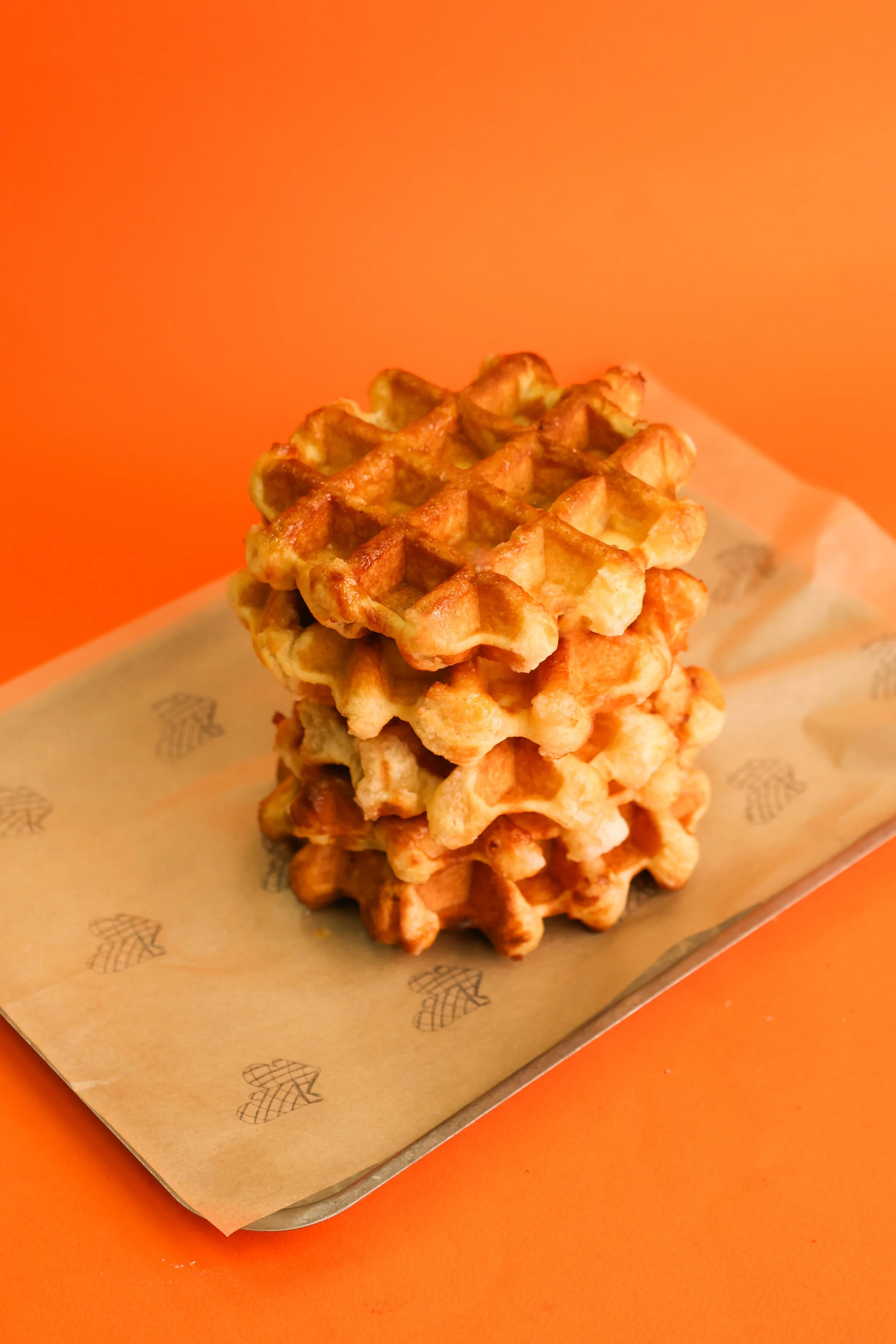 A stack of golden-brown waffles on a tray lined with parchment paper against an orange background.