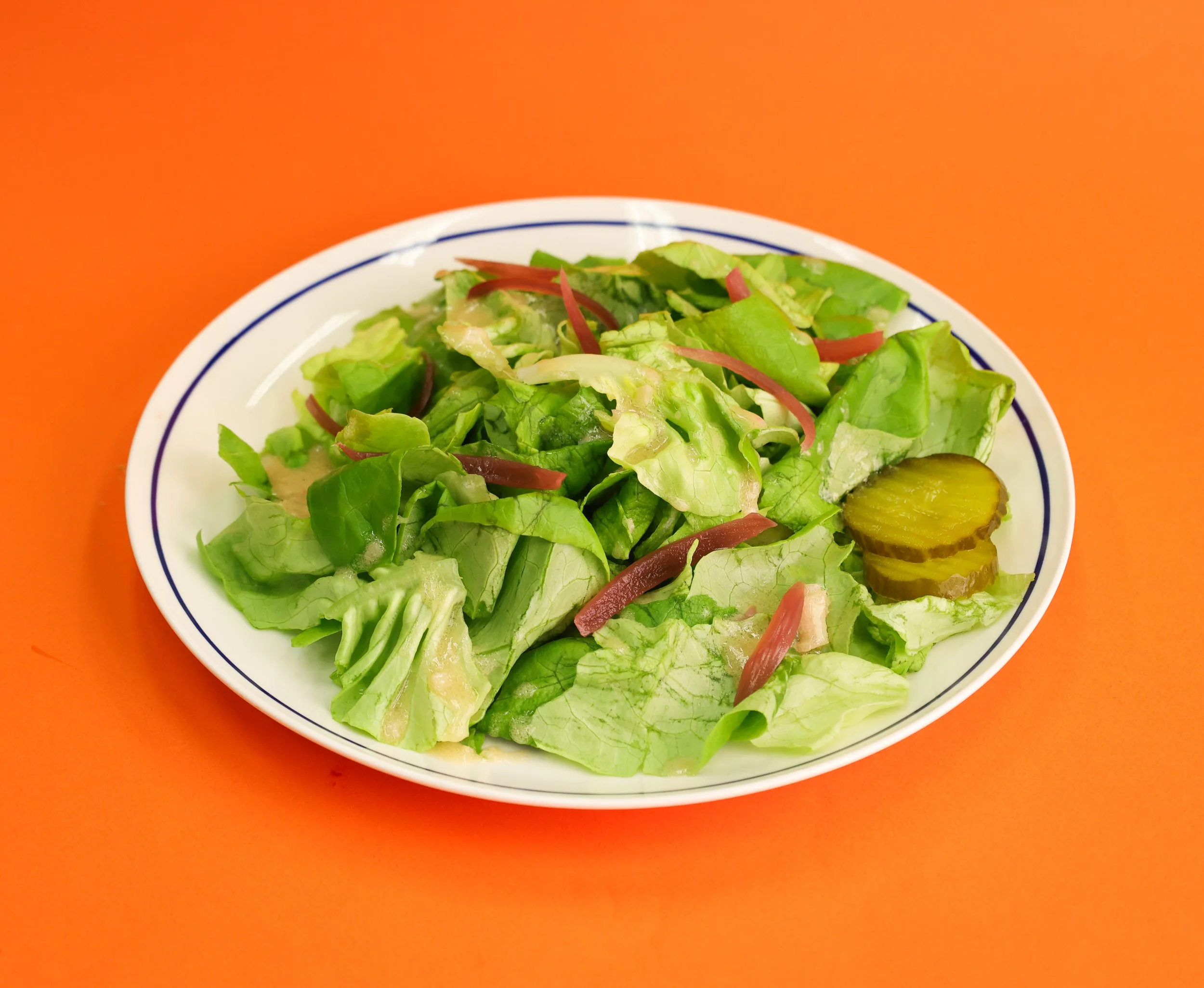 A white plate with a blue rim contains a fresh green salad with lettuce and pickles, placed on an orange surface.