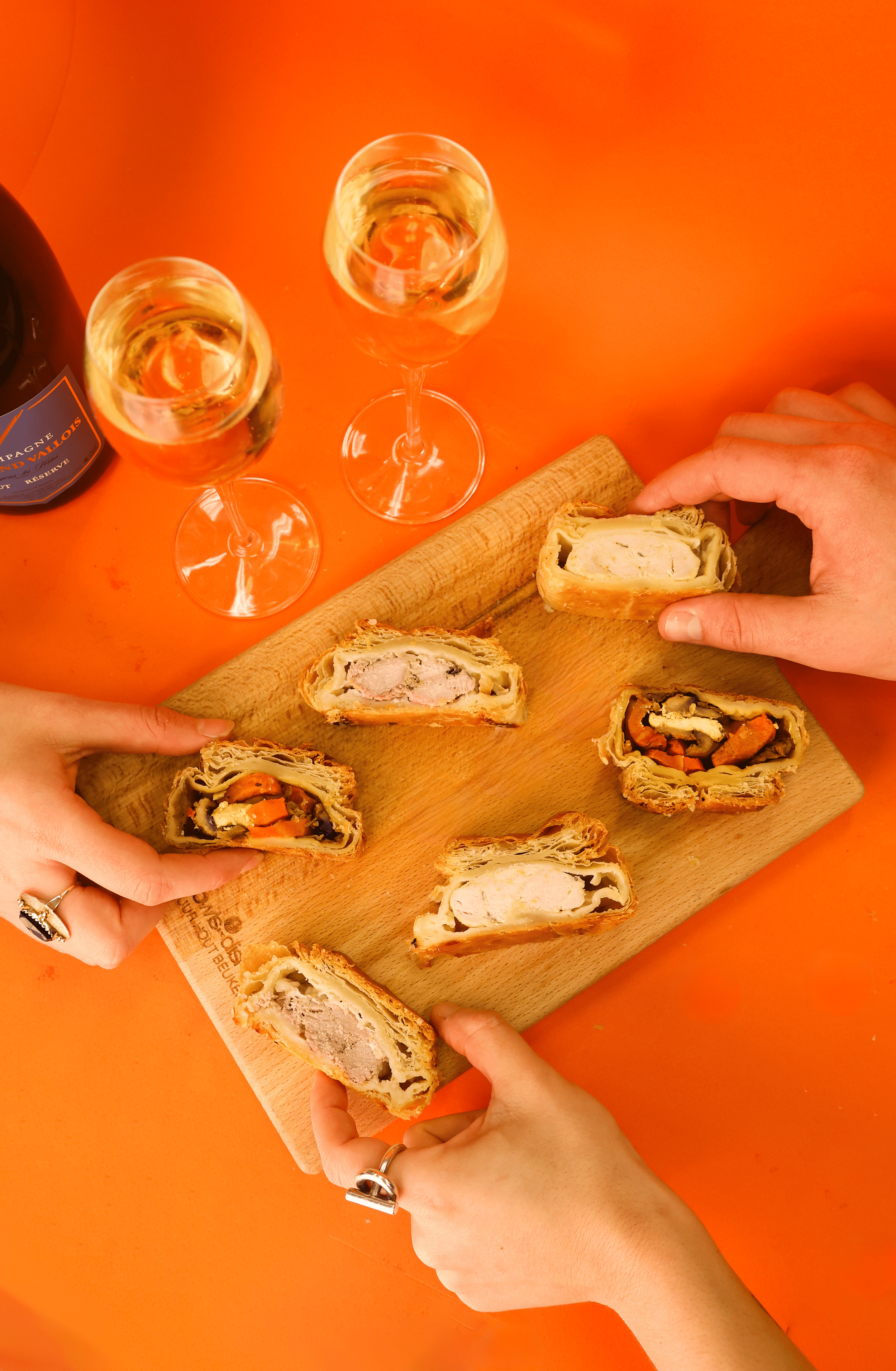 Two hands holding portions of assorted savory pastries on a wooden cutting board, with two glasses of champagne and a bottle of champagne on an orange table.