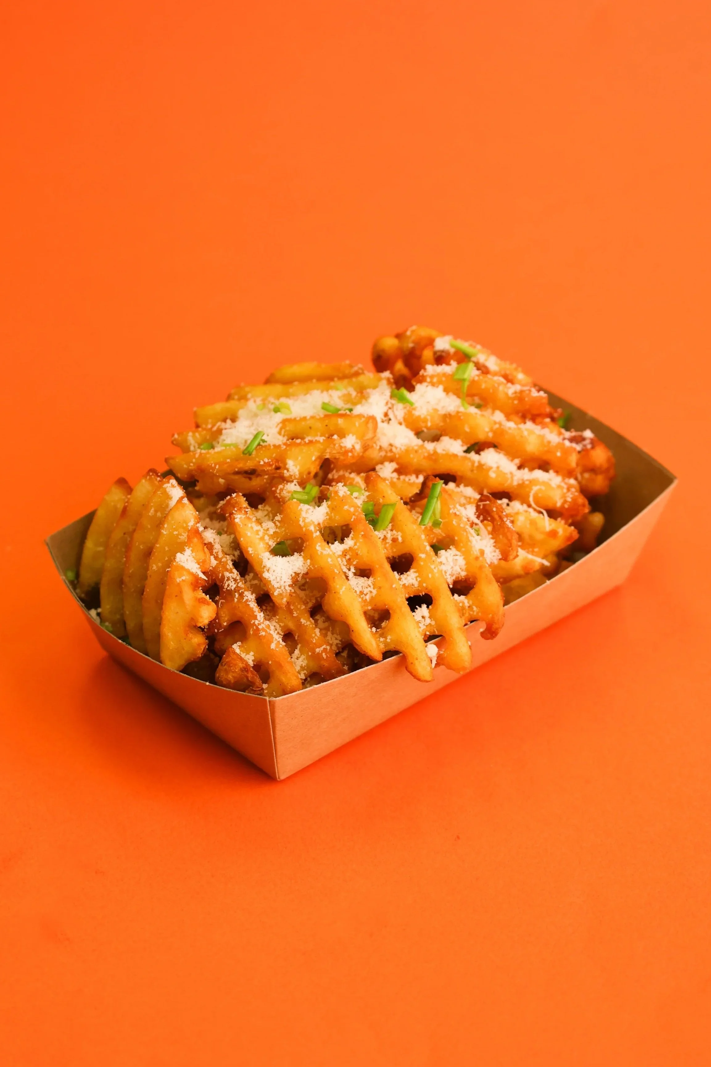 Waffle fries topped with grated cheese and chopped green herbs in a brown paper food tray on an orange background.
