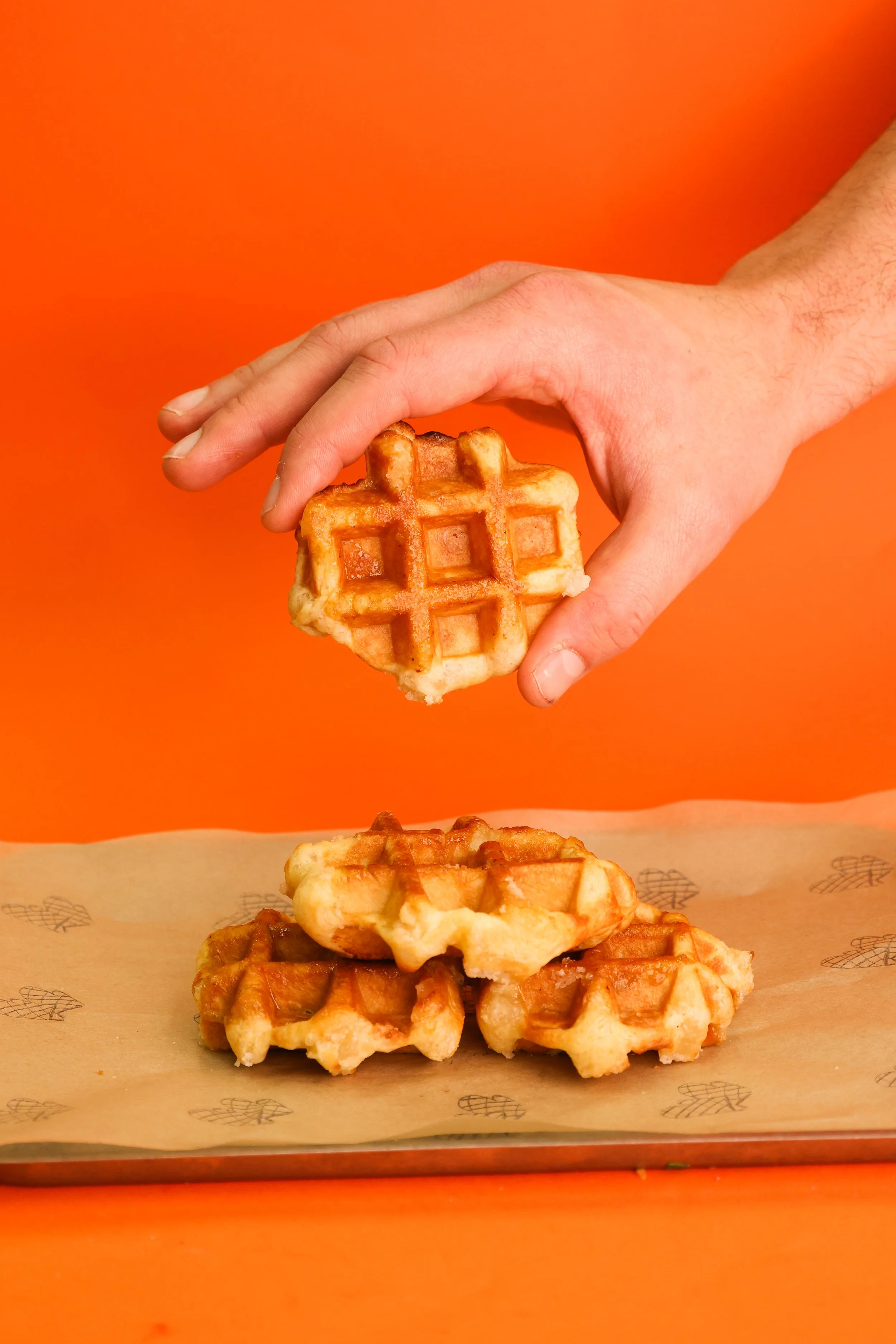 A hand holding a waffle above three stacked waffles on a tray, with an orange background.