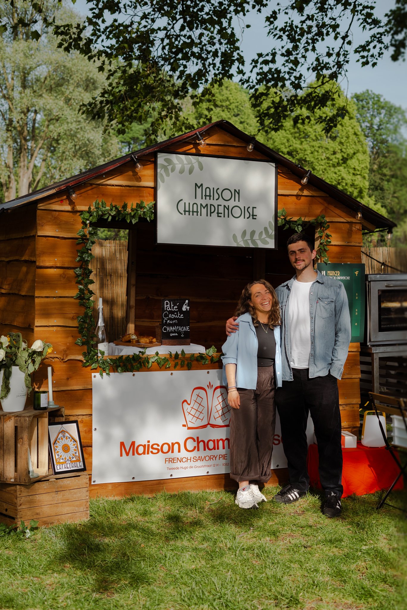 Two people standing in front of a food stall named Maison Champenoise, with a sign advertising French savory pies. The stall is made of wood and decorated with green foliage. The man has short dark hair, a beard, and is wearing a light denim jacket and black sneakers, while the woman has curly hair and is wearing a light blue jacket and gray pants. The setting appears to be outdoors during the daytime with trees and a grassy area around.