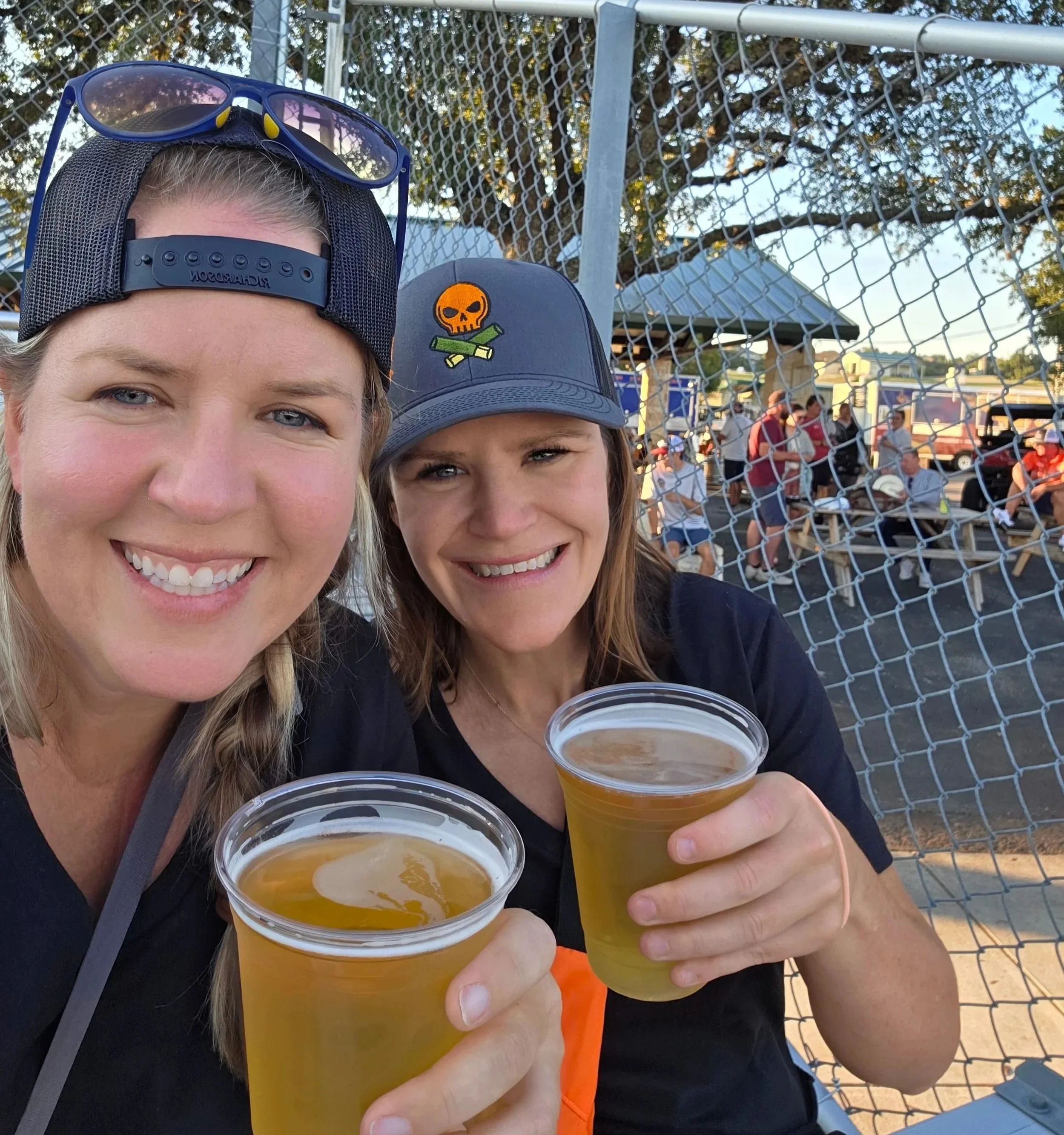Two women are smiling and holding cups of beer at an outdoor event, with a chain-link fence and a crowd in the background.