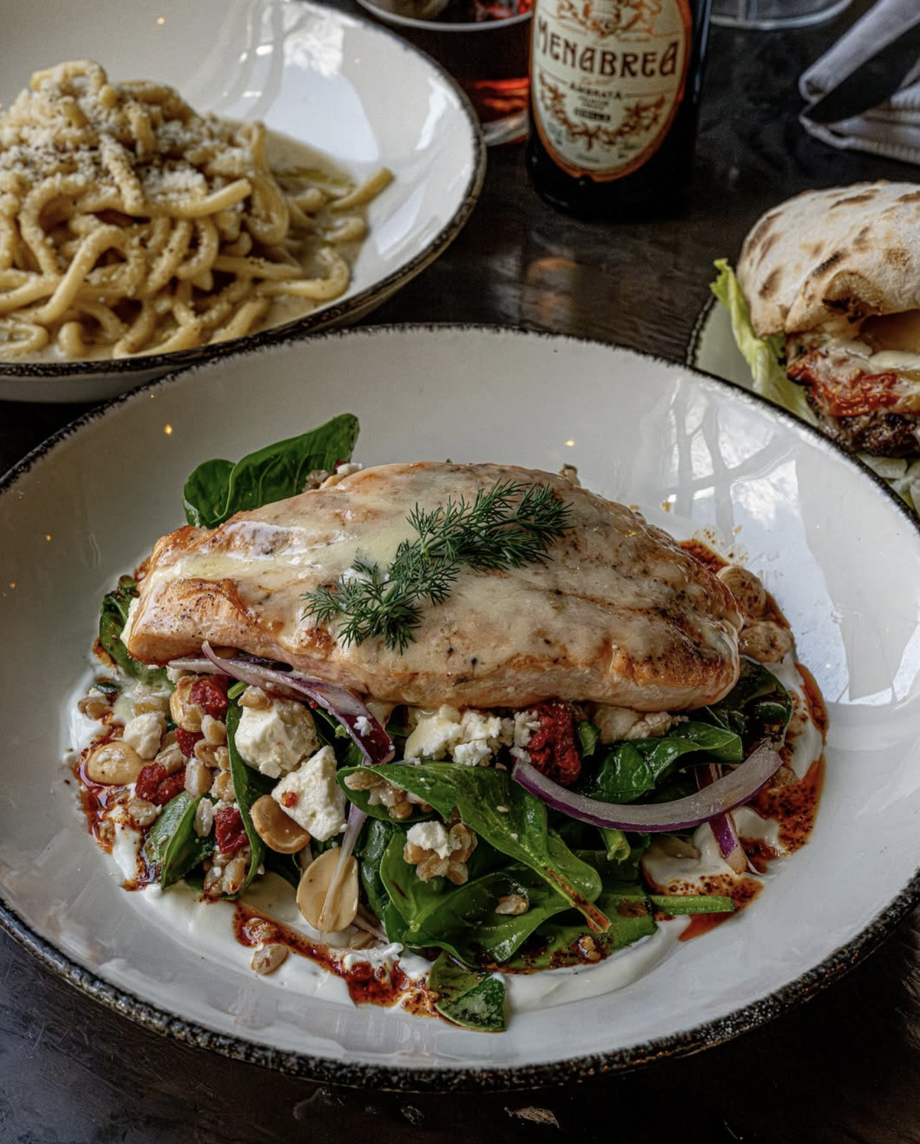 A plate of grilled chicken with herbs on top, served with a salad of greens, onions, nuts, feta cheese, and a creamy sauce. In the background, there is a bowl of pasta, a bottle of beer, and a sandwich with grilled meat, cheese, and vegetables.