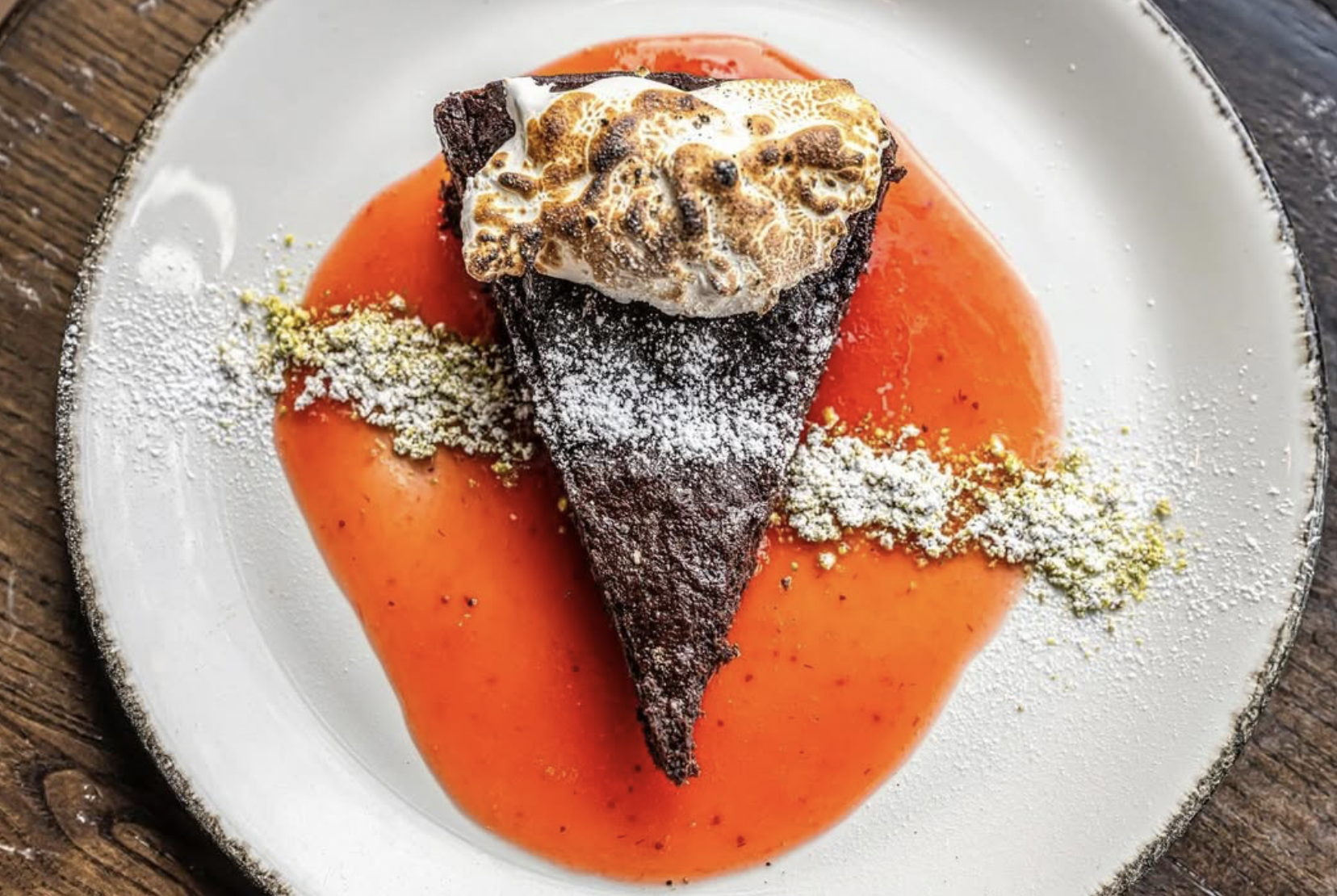 Chocolate dessert with toasted marshmallow topping on a white plate, accompanied by orange sauce and dusted with powdered sugar and green garnish.