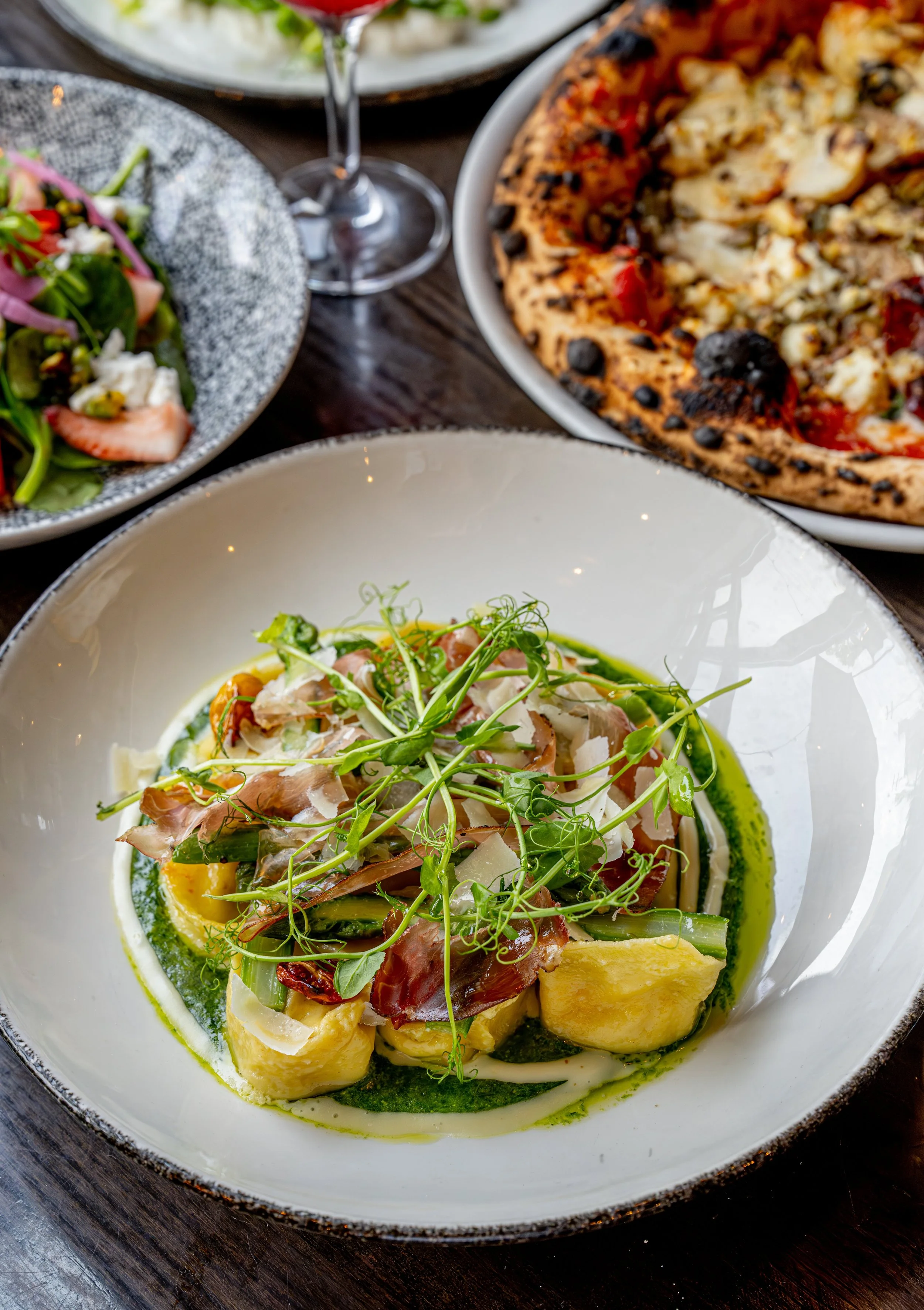 A plate of pasta garnished with microgreens, served with sauce and cheese. In the background, there is a salad, a pizza, a salad with strawberries, and a glass of red wine.