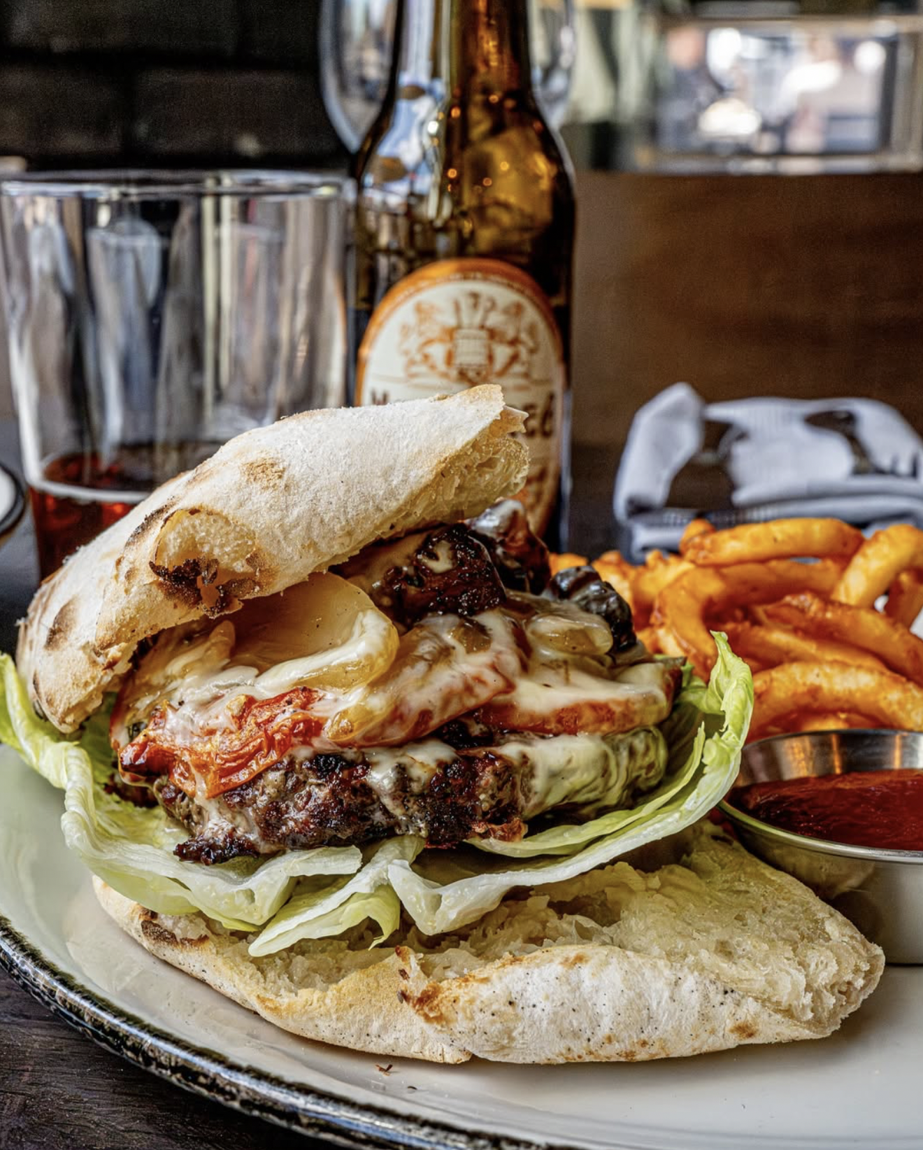 A close-up of a cheeseburger with lettuce, grilled onions, melted cheese, and tomato on a toasted bun, served with curly fries and a side of barbecue sauce, with a bottle of beer and a glass in the background.