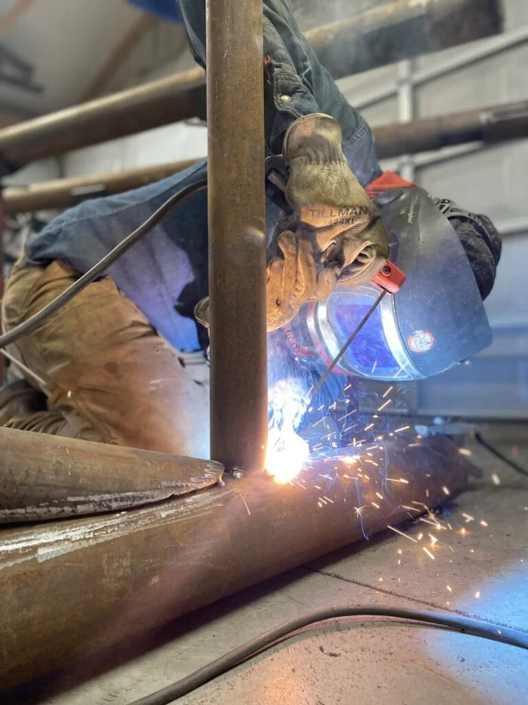 A welder wearing protective gear welding a metal pipe, with sparks flying in a workshop.