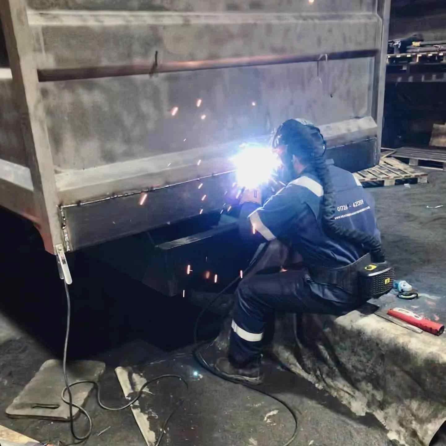 A worker welding metal on a cart outside at night, wearing safety gear including gloves and a helmet.