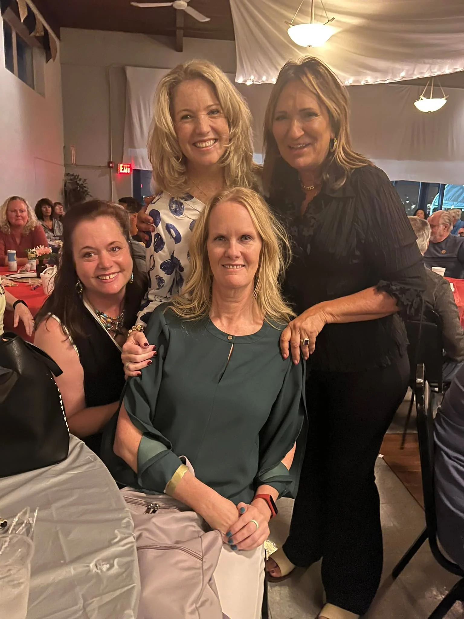 Four women at a social gathering in a restaurant, smiling for the camera. Two are seated, and two are standing behind them. The background shows other people seated at tables, with a white draped ceiling and hanging lights.