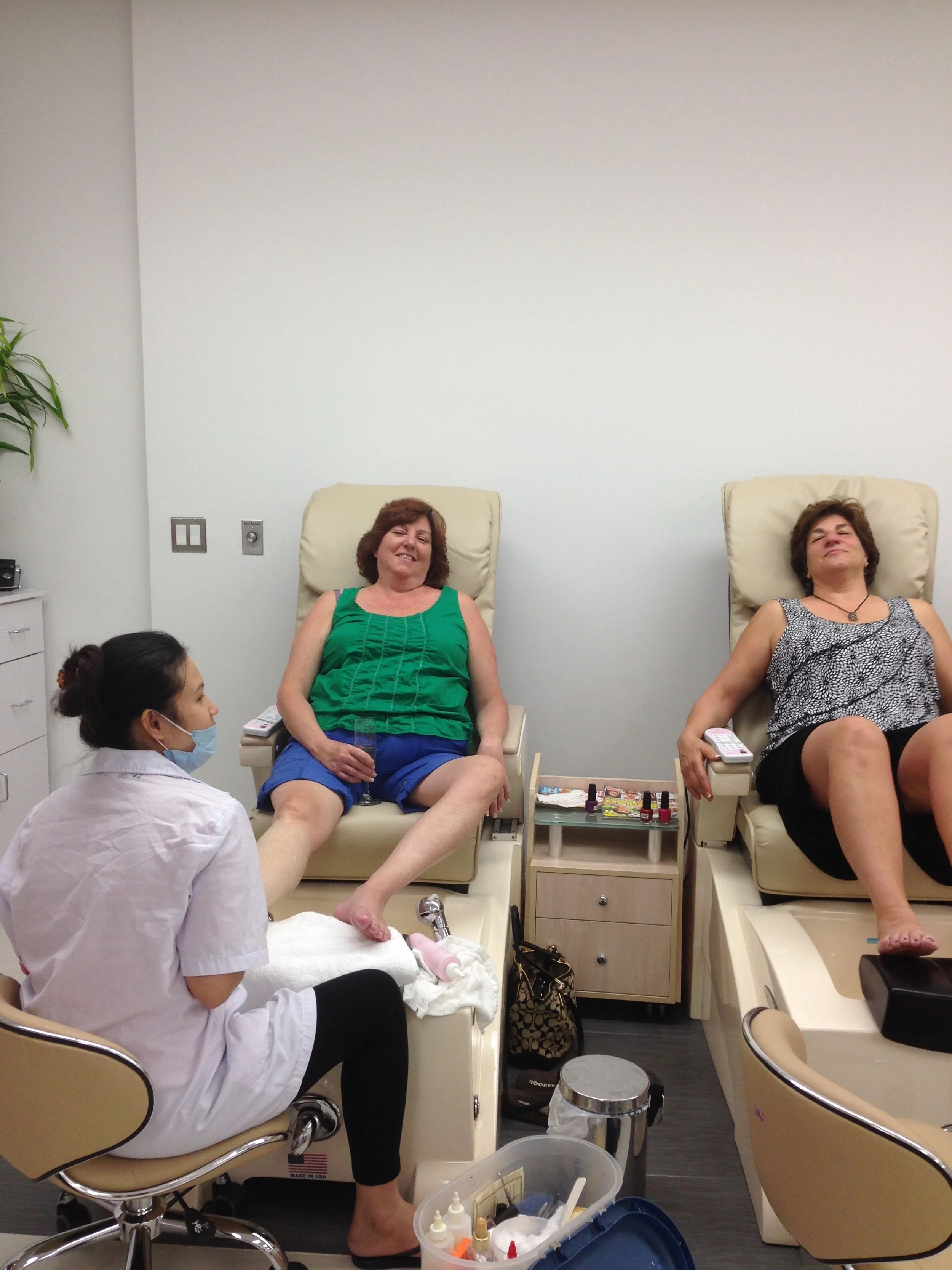 Two women sitting in reclining chairs in a nail salon, with a technician attending to one of them.