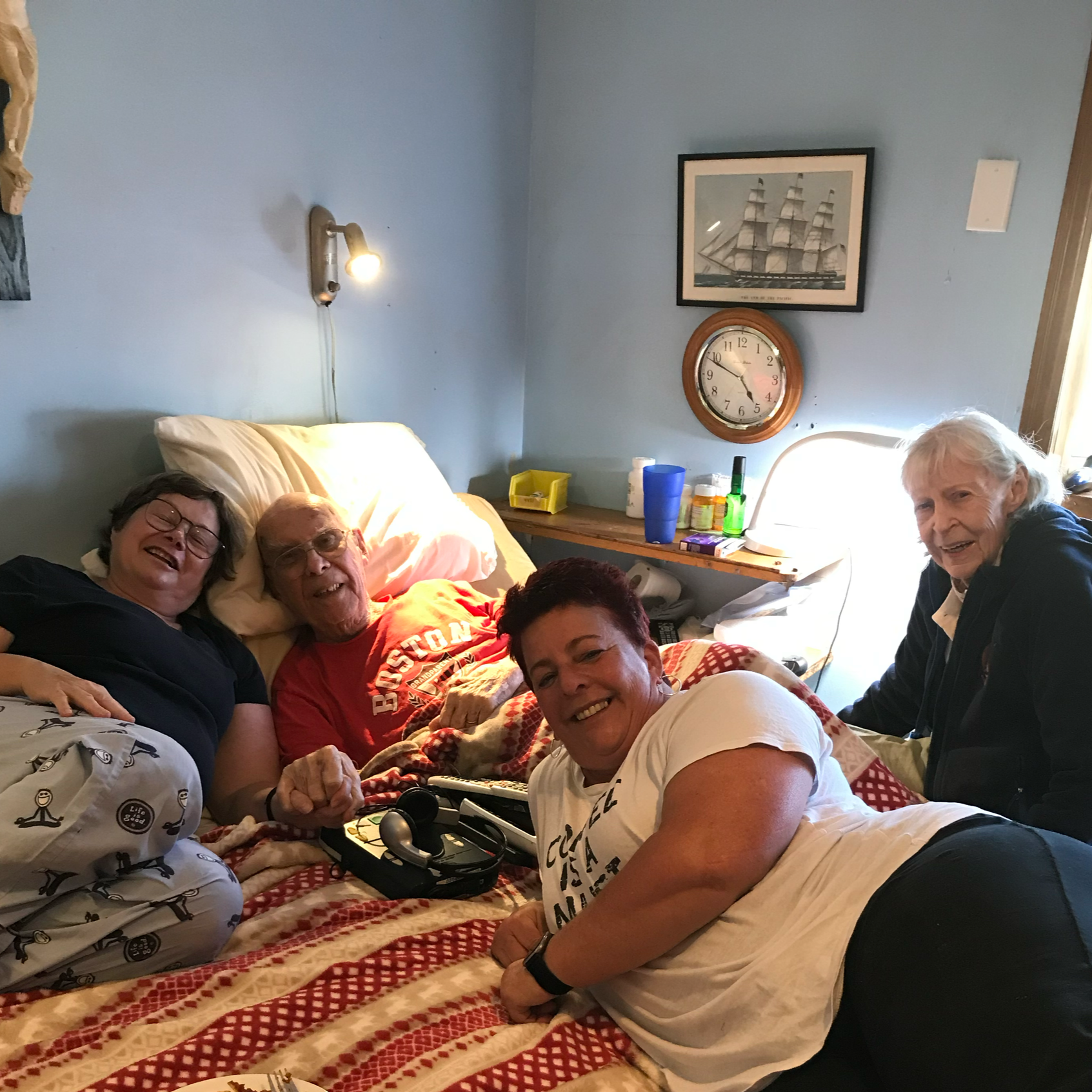 Four people gathered in a bedroom, smiling and enjoying each other's company. One person is lying in bed with a red T-shirt, while three others are leaning on the bed or sitting nearby. The room has a blue wall, a clock, and framed picture of a ship.