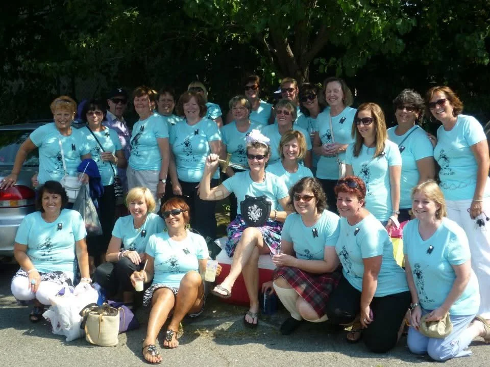 Group of women wearing matching light blue T-shirts, gathered outdoors under a tree, some sitting and some standing, smiling for the photo during daytime.