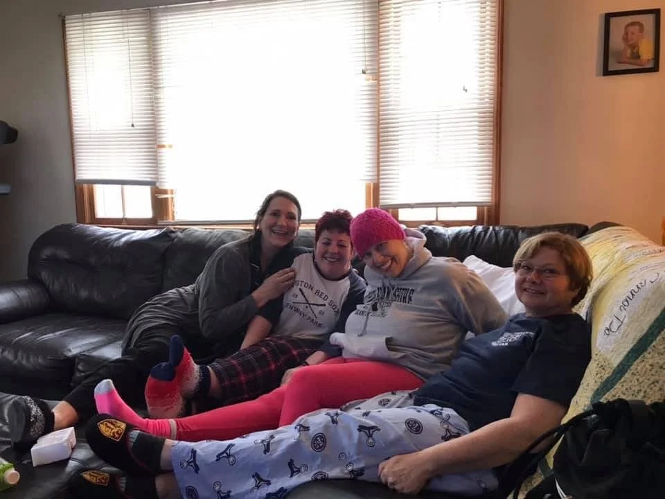 Four people sitting on a couch in a living room, smiling and enjoying each other's company, with a window with blinds behind them.