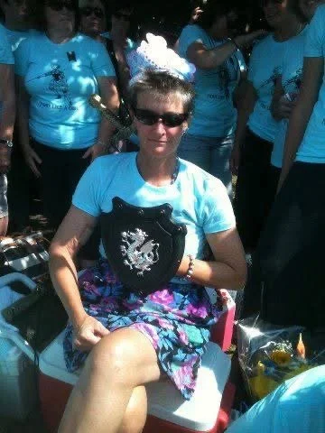 Woman with short hair, wearing sunglasses, a light blue T-shirt, a tiara, and a floral skirt, sitting outdoors surrounded by a group of people.