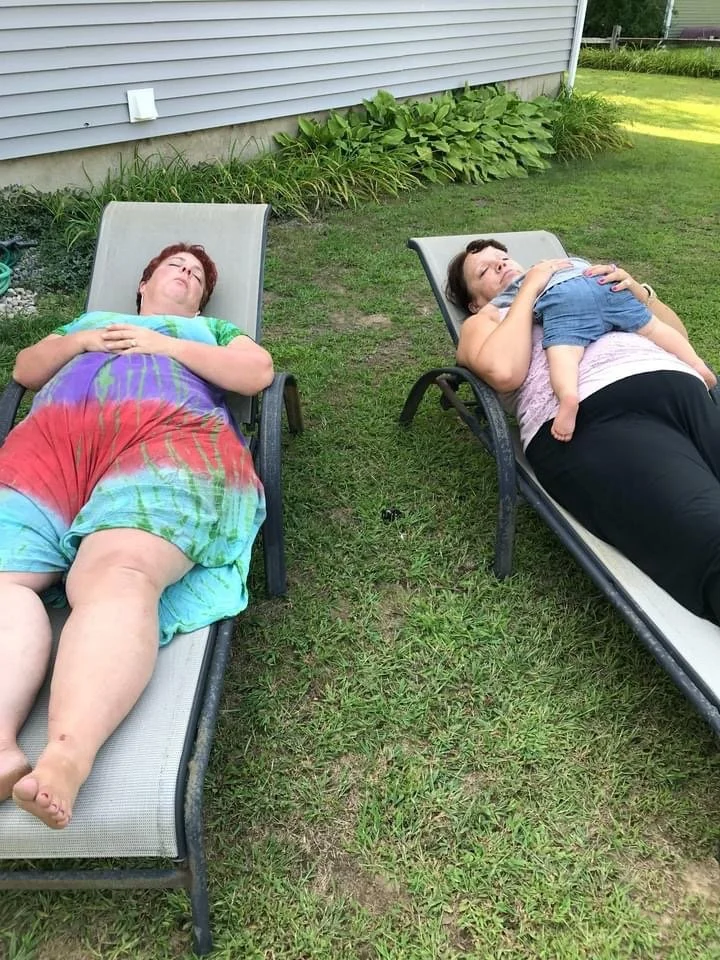 Two women are lying on lounge chairs outdoors on grass, sunbathing with their eyes closed.
