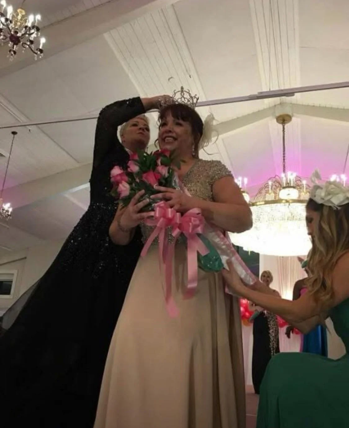 A woman is being crowned at a celebration, holding a bouquet of pink roses with pink ribbons, while another woman is helping her.