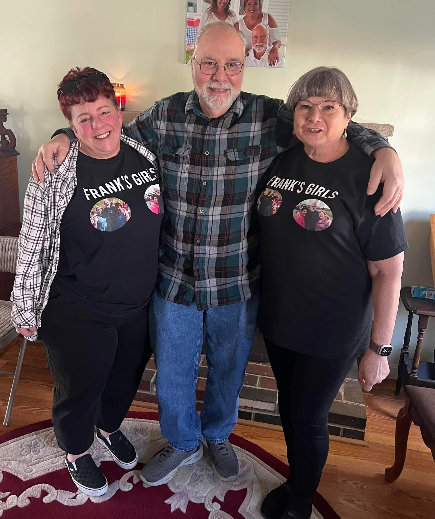 Three people standing close together indoors, smiling. Mostly cloudy wall with photo on it in background. The two women wear matching black T-shirts with a photo and text that reads 'Frank's Girls,' and the man in the middle wears a plaid shirt.