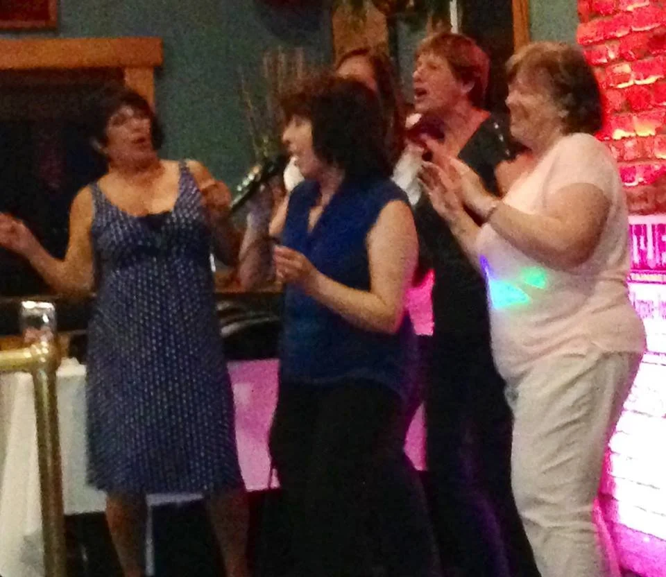 Five women are gathered at a social event, engaging in conversation and laughter, with a brick wall and colorful lighting in the background.