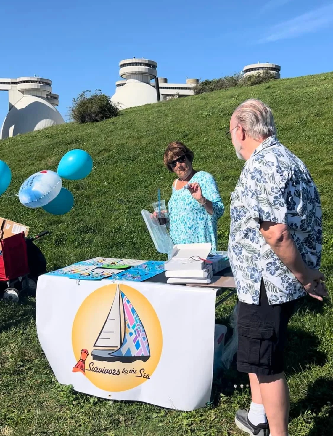 A man and a woman standing next to a table with a banner reading 'Survivors by the Sea' on a grassy hill. The woman is holding a drink and gesturing with her hand, while the man, with his back to the camera, is engaged in conversation. There are blue