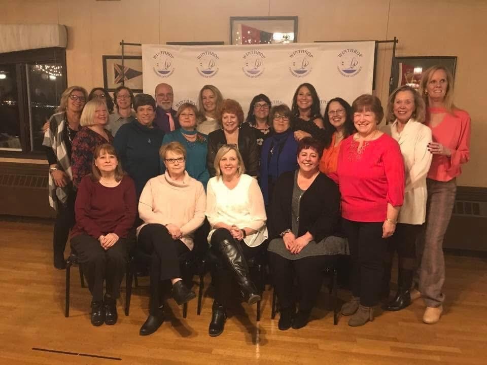 A group of women gathered together for a photo at an indoor event, with a white banner in the background featuring a logo with a stylized sailboat and the word 'SYNTHROP'.