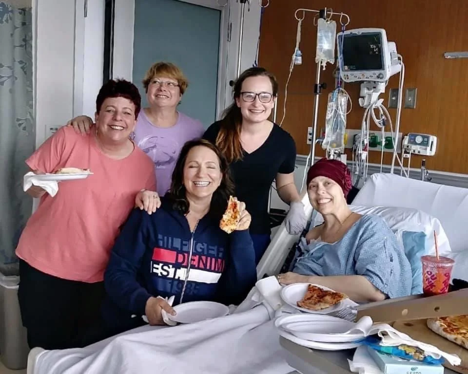 Group of five women celebrating in a hospital room with a patient in bed, holding pizza slices.
