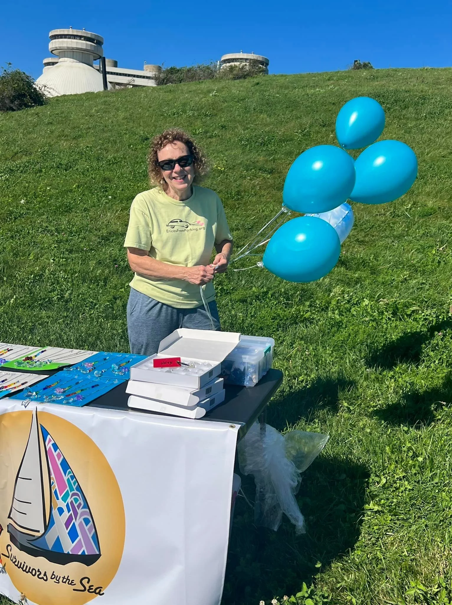 Woman standing outdoors at a booth with colorful jewelry, holding blue balloons, with a grassy hill and a large modern building in the background.
