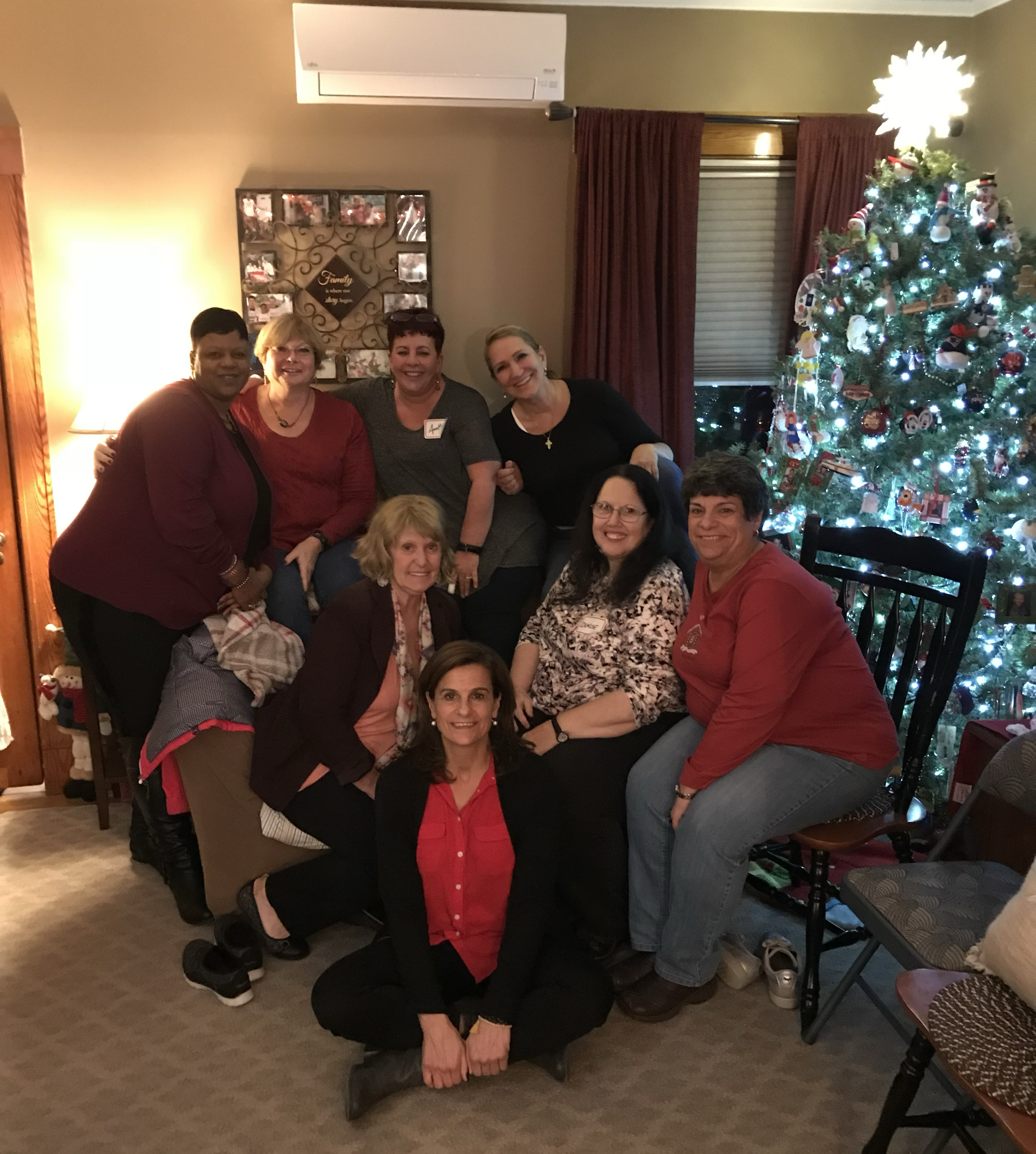 A group of nine women gathered in a living room decorated for Christmas with a lit Christmas tree and a holiday family photo display on the wall.