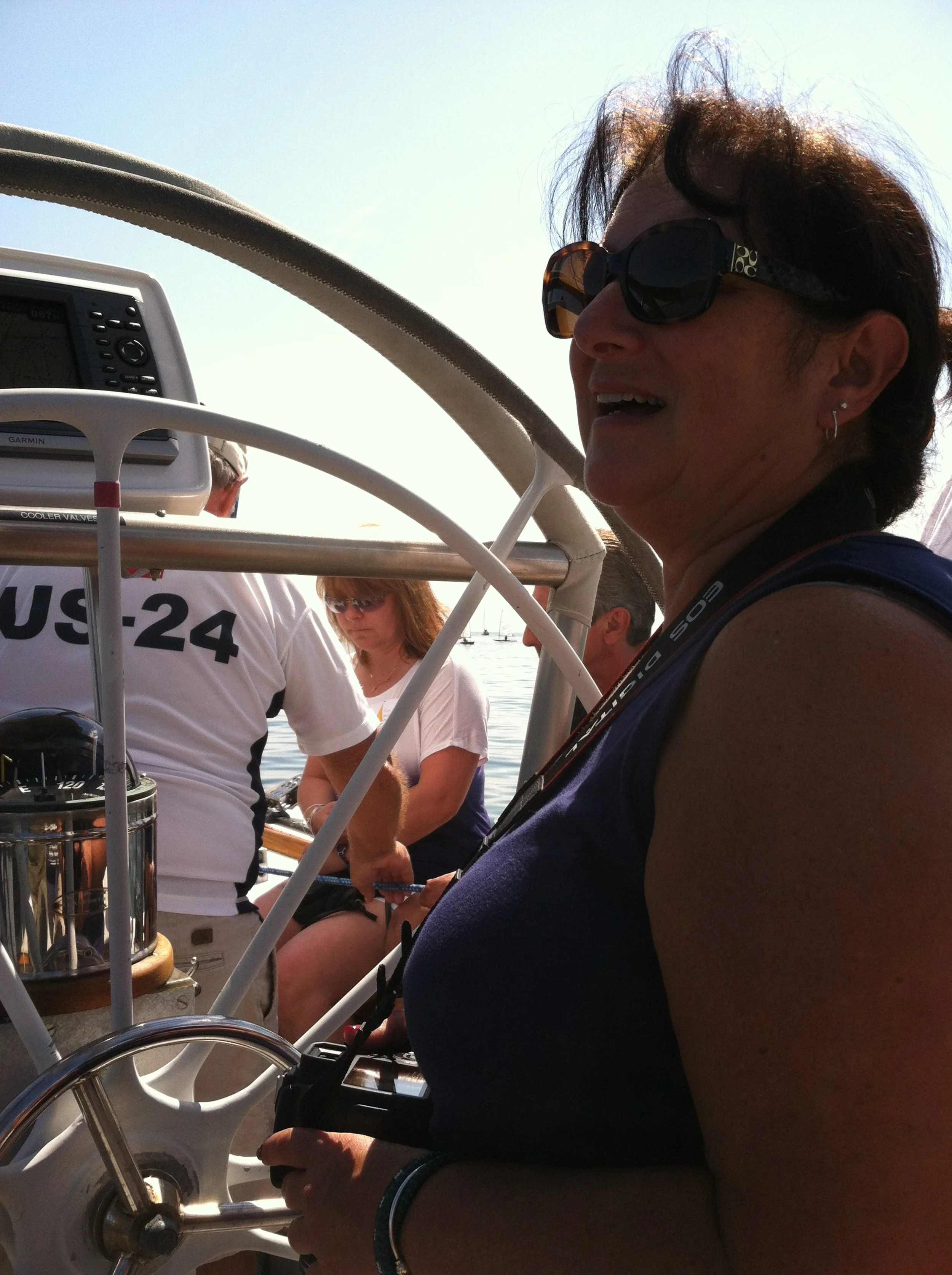 A woman wearing sunglasses and a sleeveless top, smiling on a boat with a steering wheel and navigation equipment, with other people in the background near the water.