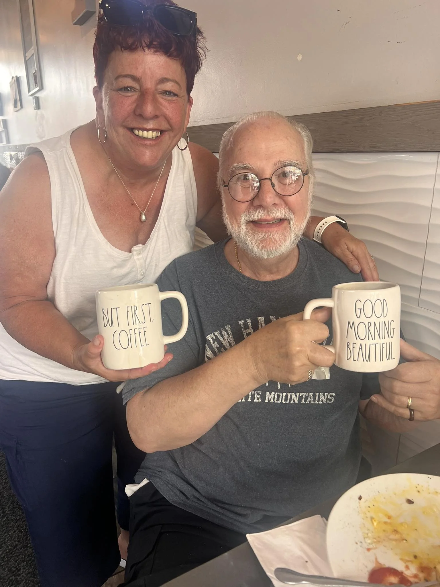 Two women and a man sitting at a table, smiling and holding coffee mugs. The woman standing is wearing a white tank top and sunglasses on her head, holding a mug that reads 'BUT FIRST, COFFEE'. The man sitting is wearing a gray T-shirt, glasses, and 