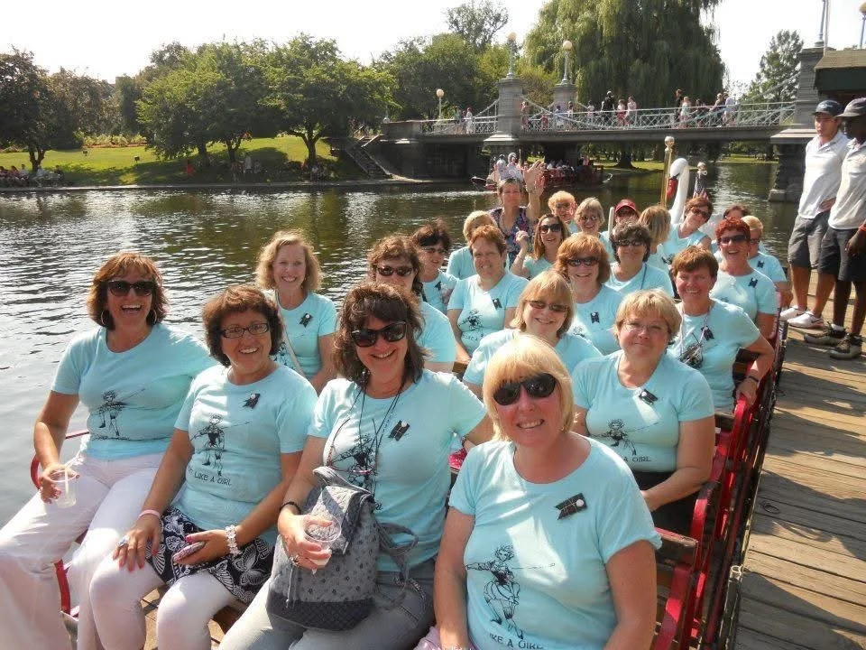 A group of women wearing matching light blue T-shirts sitting on a boat on a river, smiling and enjoying a sunny day.