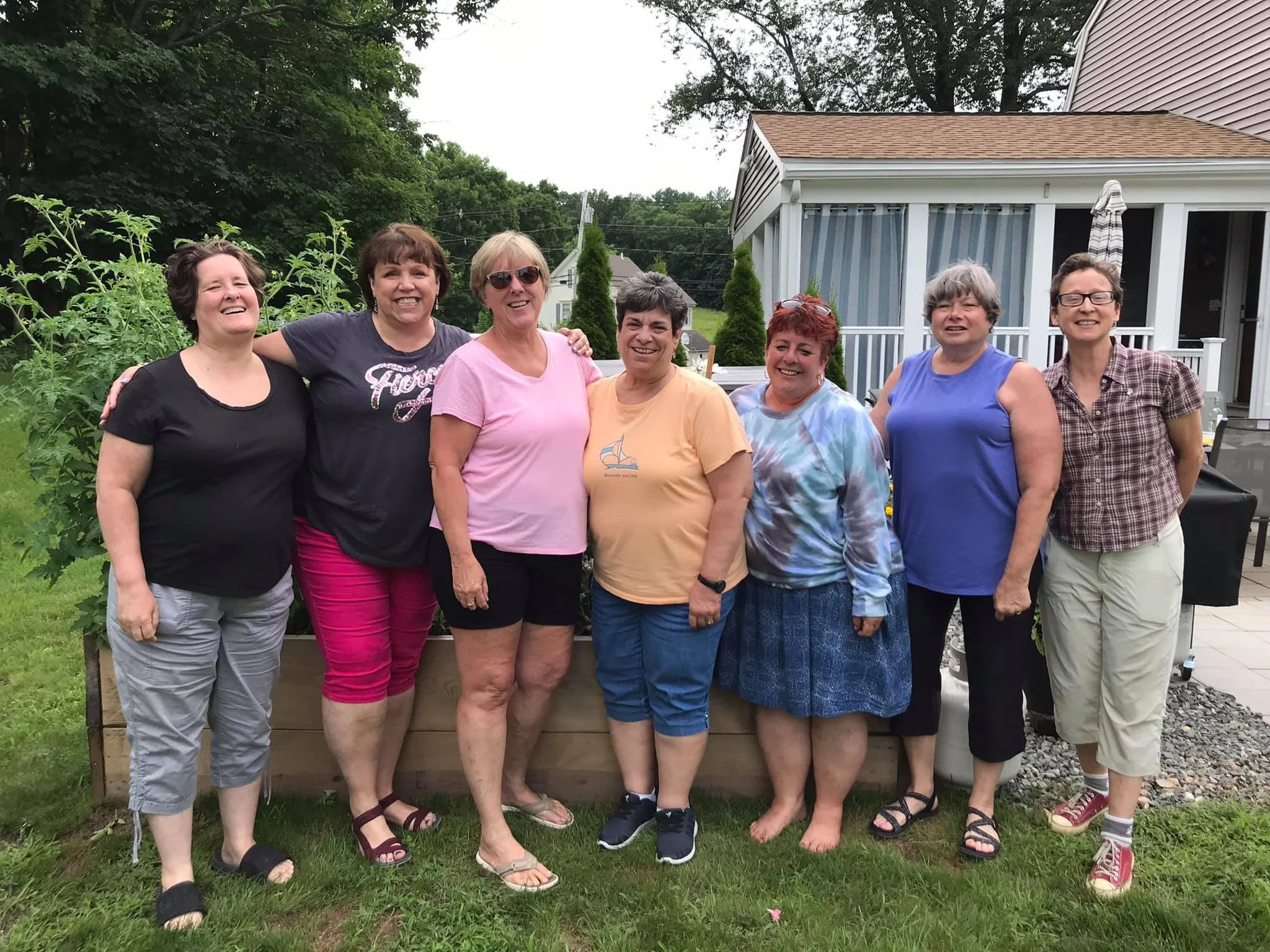 Group of eight women standing outdoors in a backyard, smiling for a photo, with a house and trees in the background.
