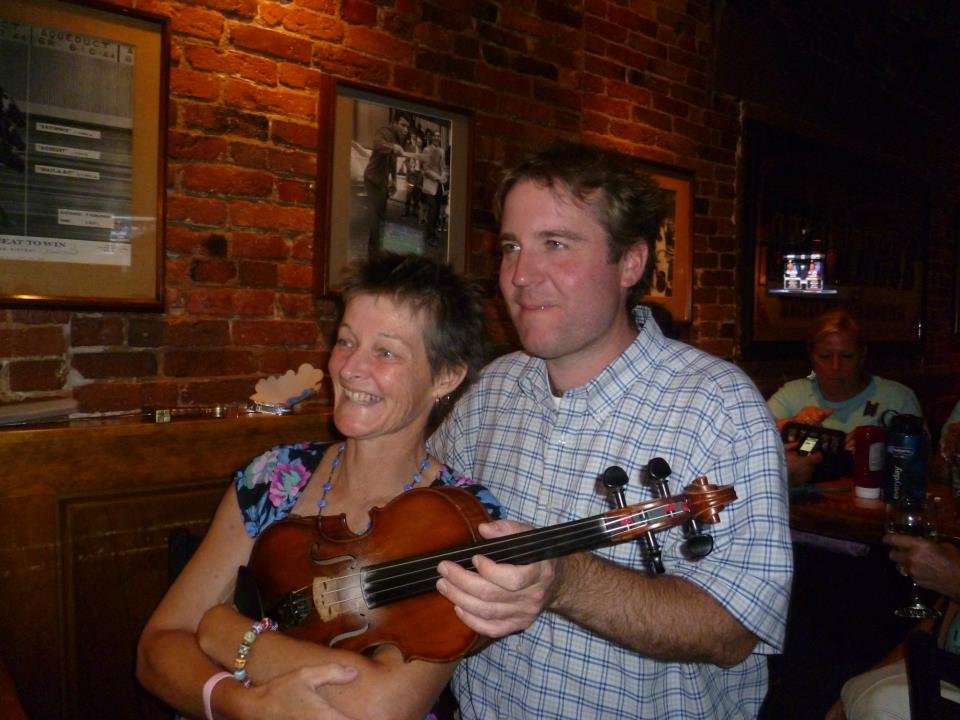 A woman smiling and holding a violin, standing next to a man in a checkered shirt in a dimly-lit brick-walled venue.