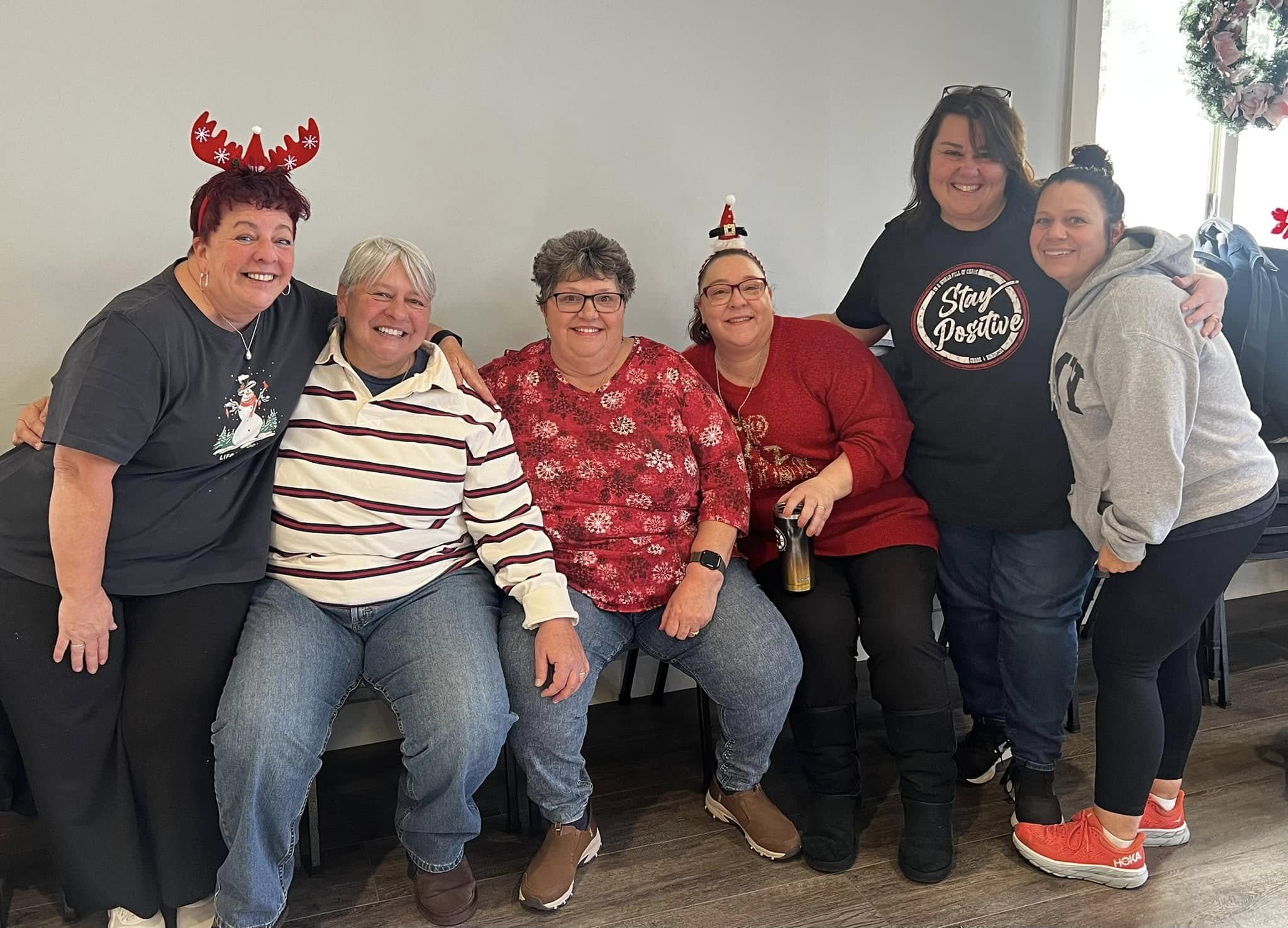 Group of six women wearing holiday-themed clothing and accessories, smiling indoors, decorated for Christmas.