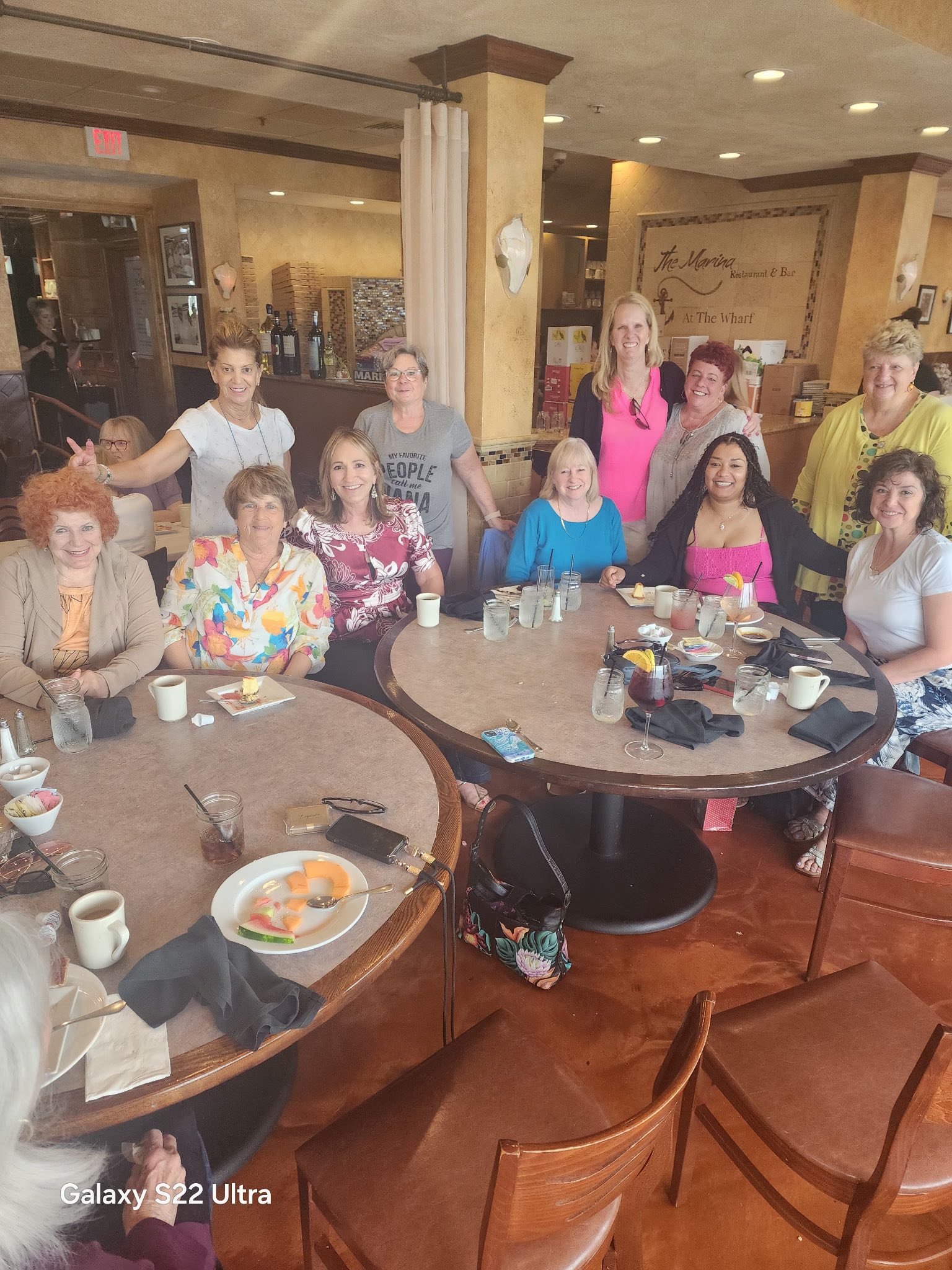 A group of women gathered around a large round table at a restaurant, smiling for the camera, with some sitting and others standing behind.