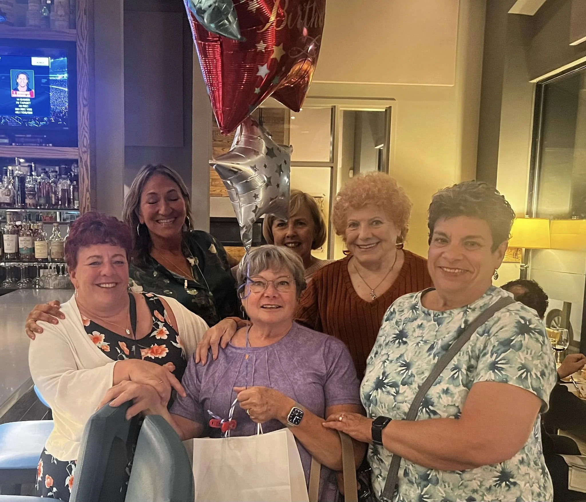 Group of six women celebrating at a restaurant, with balloons and party decorations in the background.