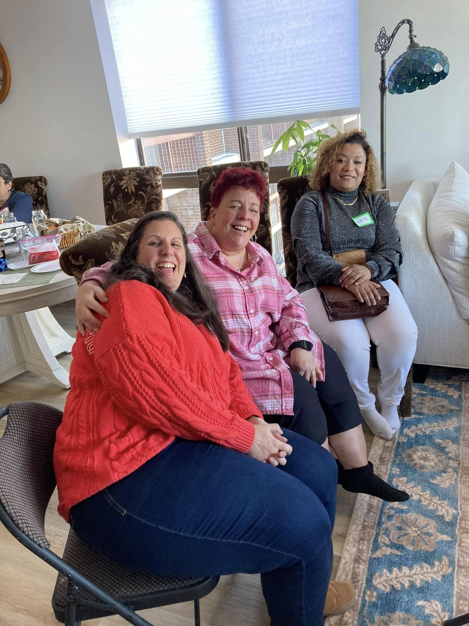 Three women sitting indoors, smiling and enjoying each other's company, in a cozy room with a window, plants, and a table in the background.