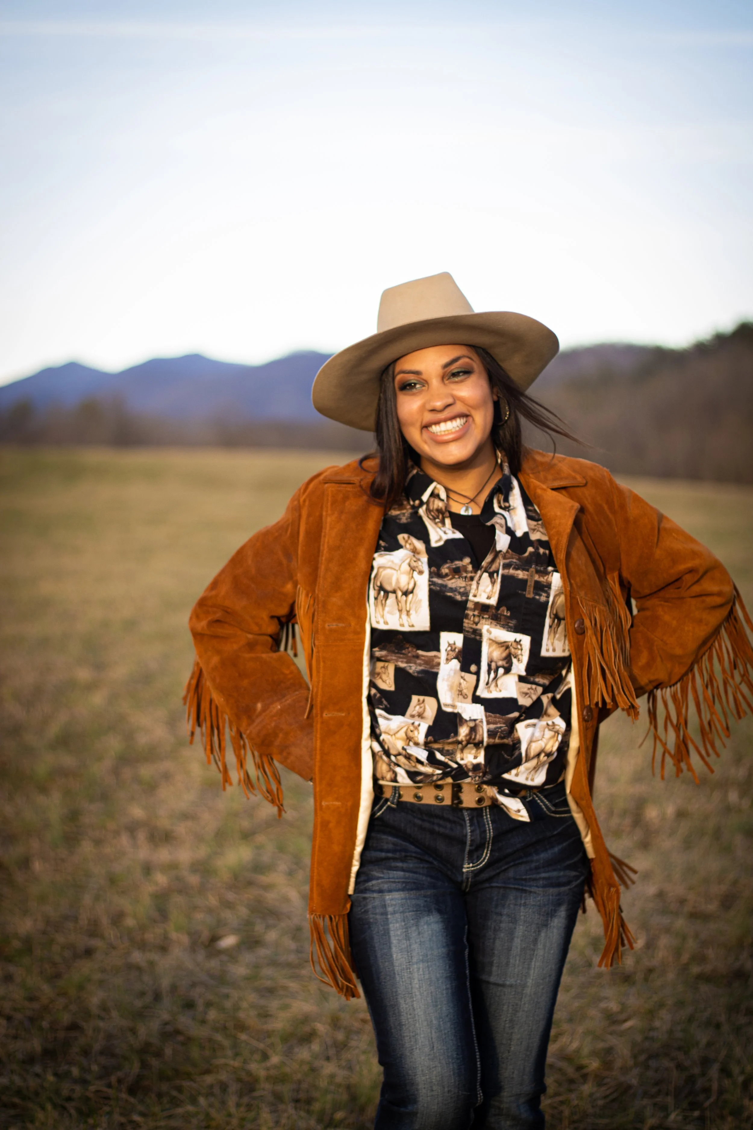 A woman standing outdoors in a field, smiling, wearing a tan cowboy hat, a brown fringe suede jacket, a graphic shirt with horse patterns, and blue jeans.