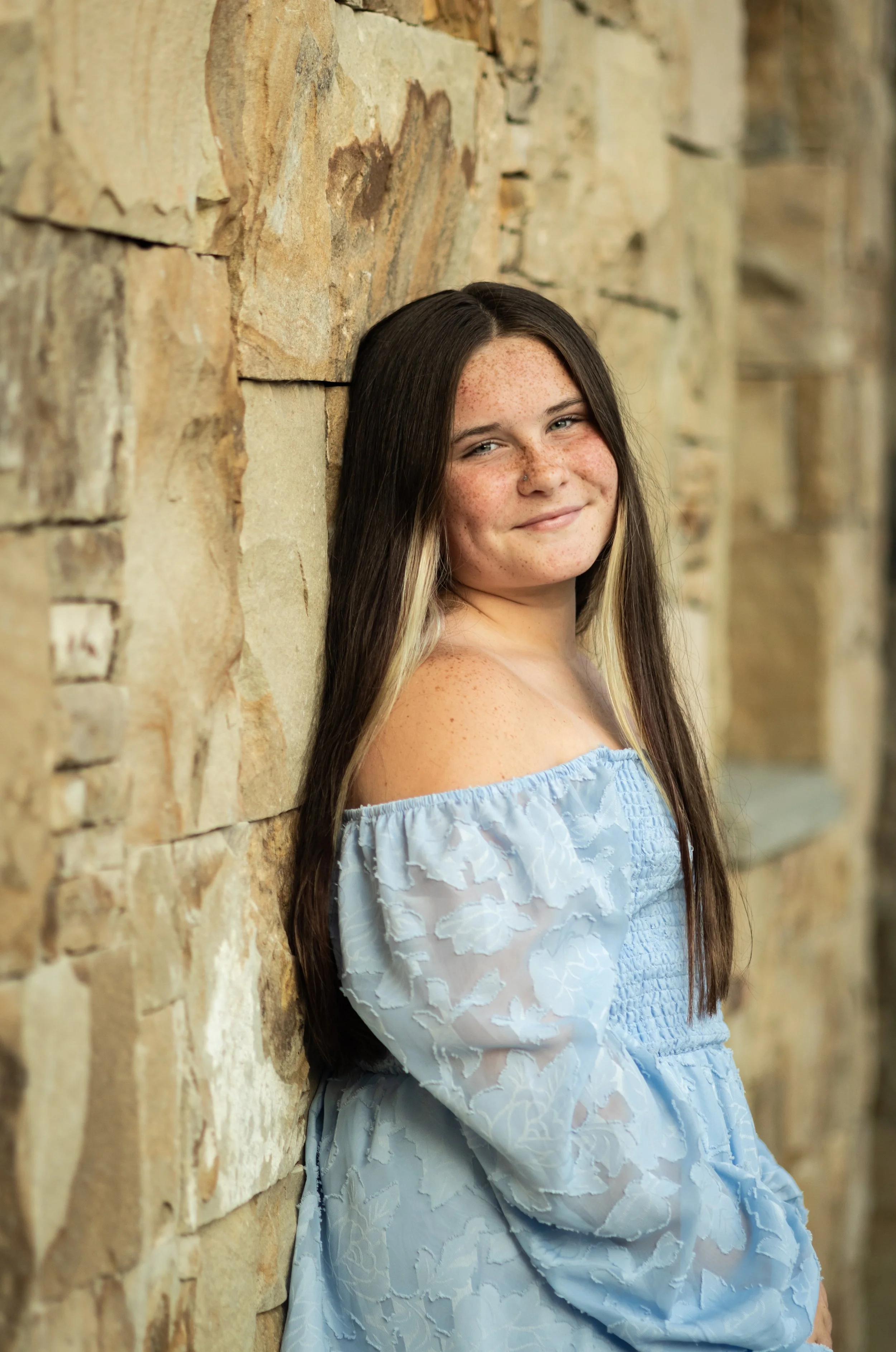 A girl with long dark hair and freckles wearing a light blue off-the-shoulder dress, leaning against a stone wall and smiling.
