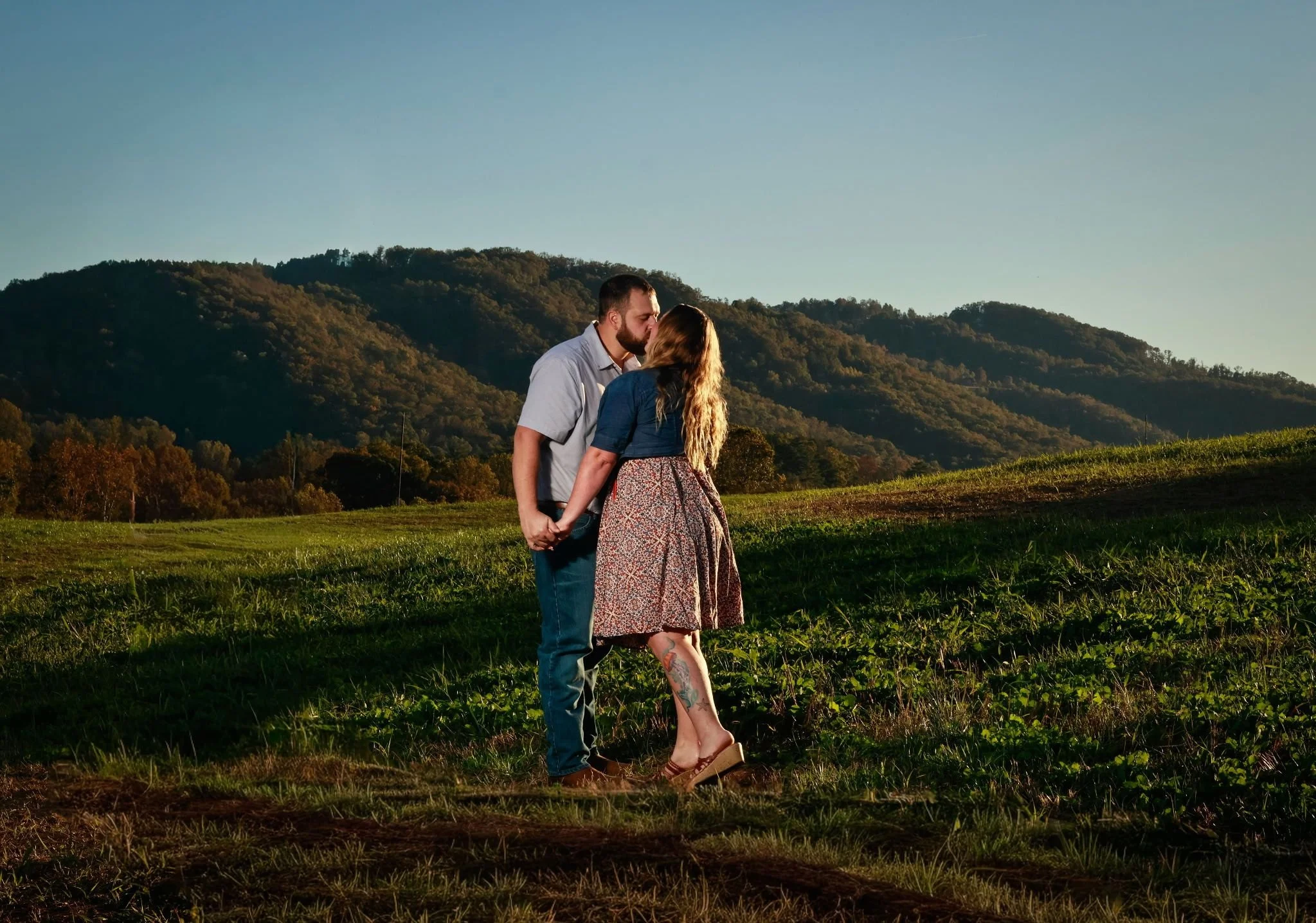 A couple holding hands and kissing outdoors on a grassy hill with mountains in the background at sunset.
