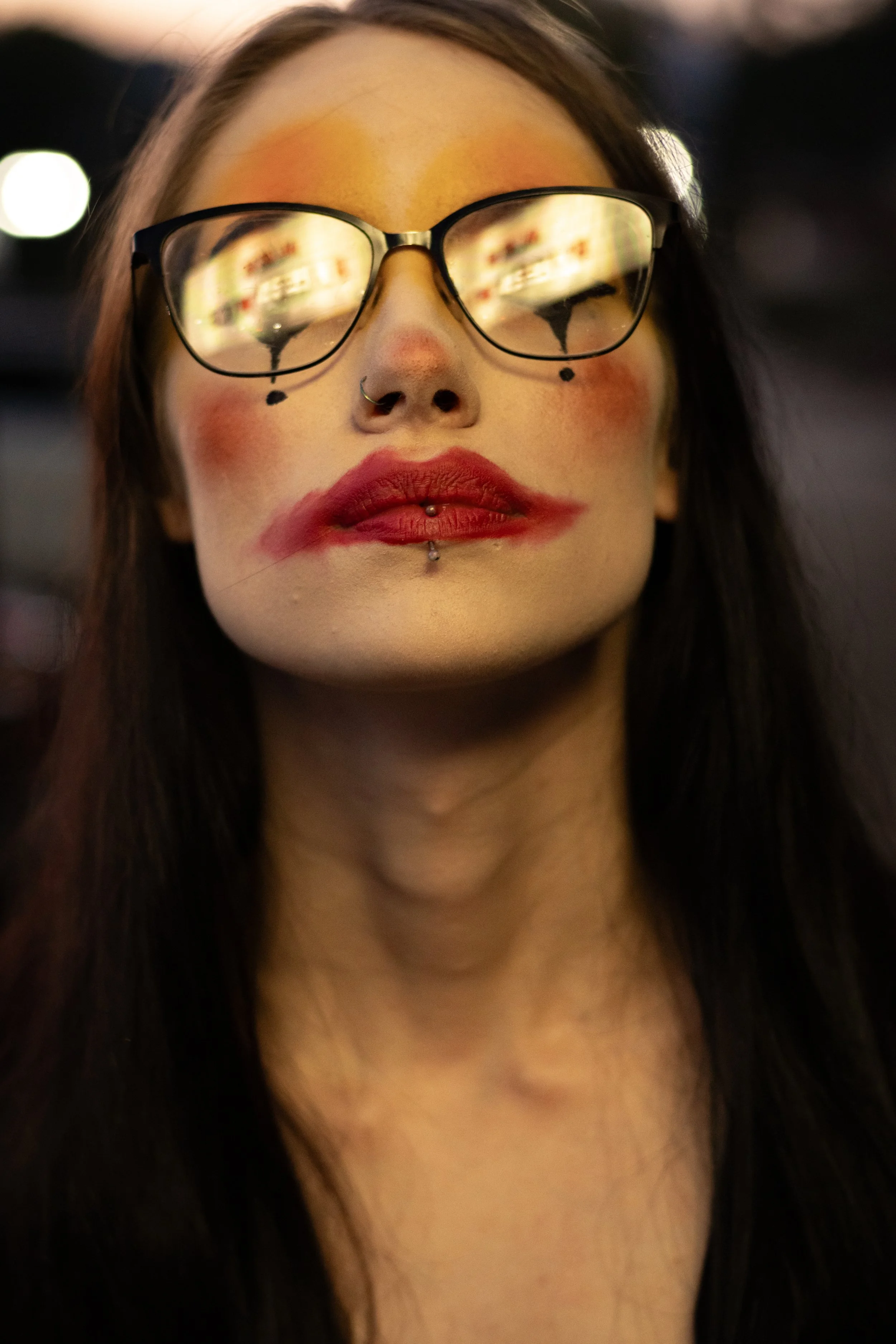 A woman with glasses, face makeup, a nose ring, and lip piercings, looking directly at the camera.