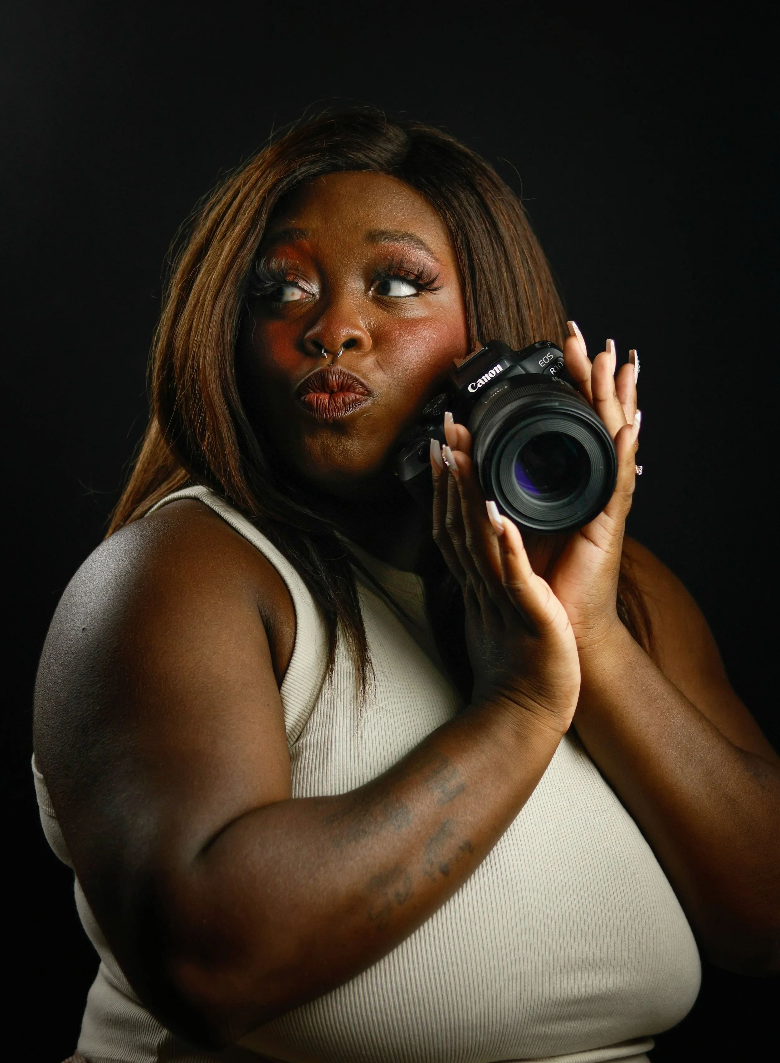 A woman with brown hair holding a Canon camera against a black background, looking off to the side with a playful expression.