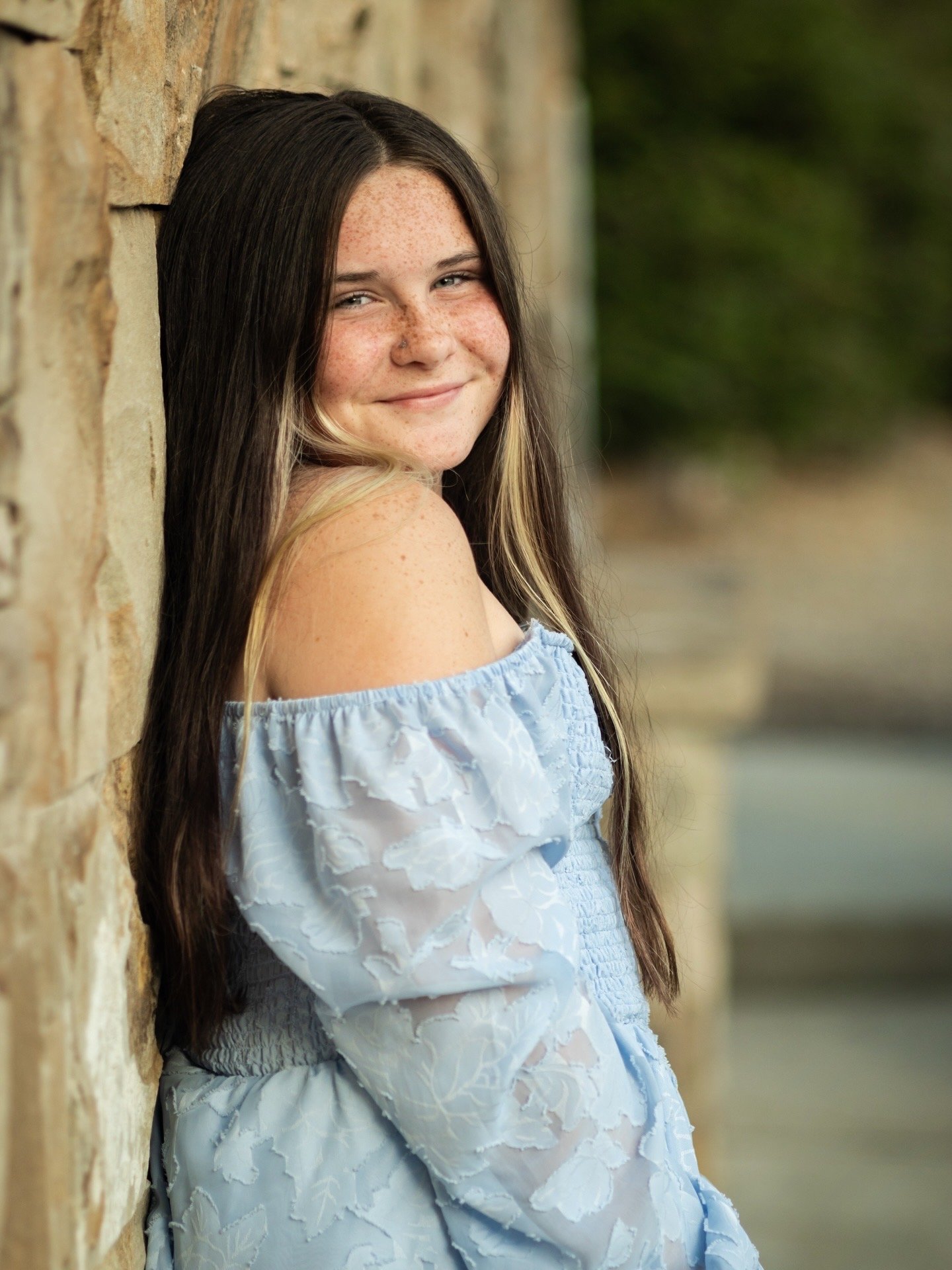 A young girl with long dark hair and freckles, smiling while leaning against a stone wall, wearing a light blue off-the-shoulder dress.