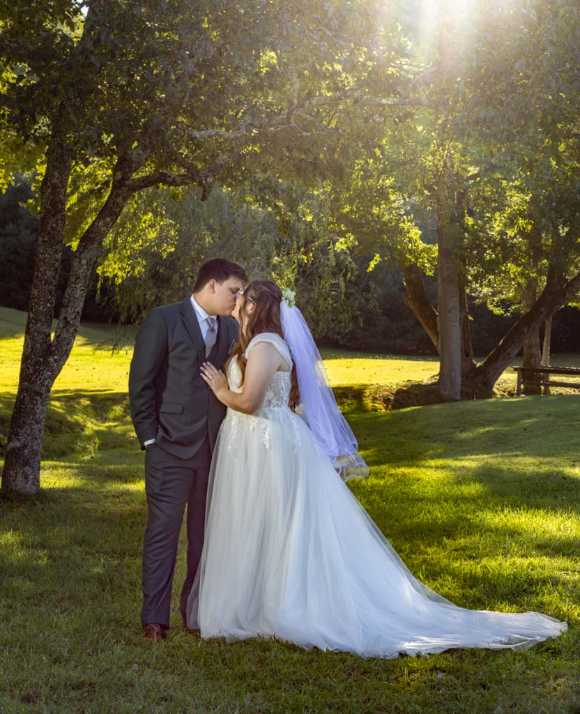 A bride and groom sharing a kiss outdoors during their wedding, surrounded by trees and sunlight.