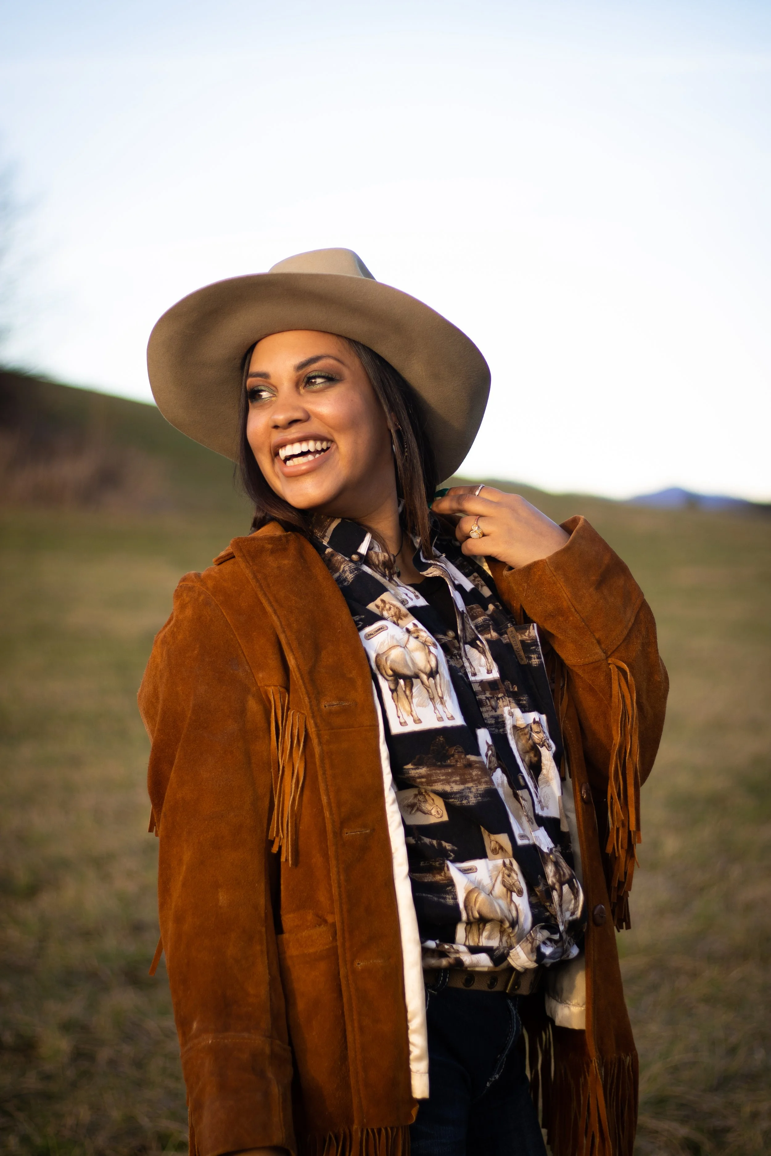 A woman wearing a cowboy hat, a patterned button-up shirt with horses, and a brown fringed suede jacket, smiling outdoors with a blurred grassy landscape in the background.