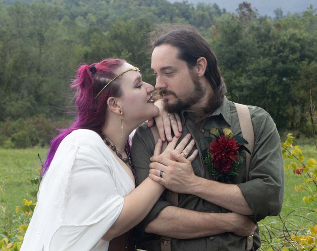 A couple with colorful hair and tattoos sharing a close moment outdoors in a green field.