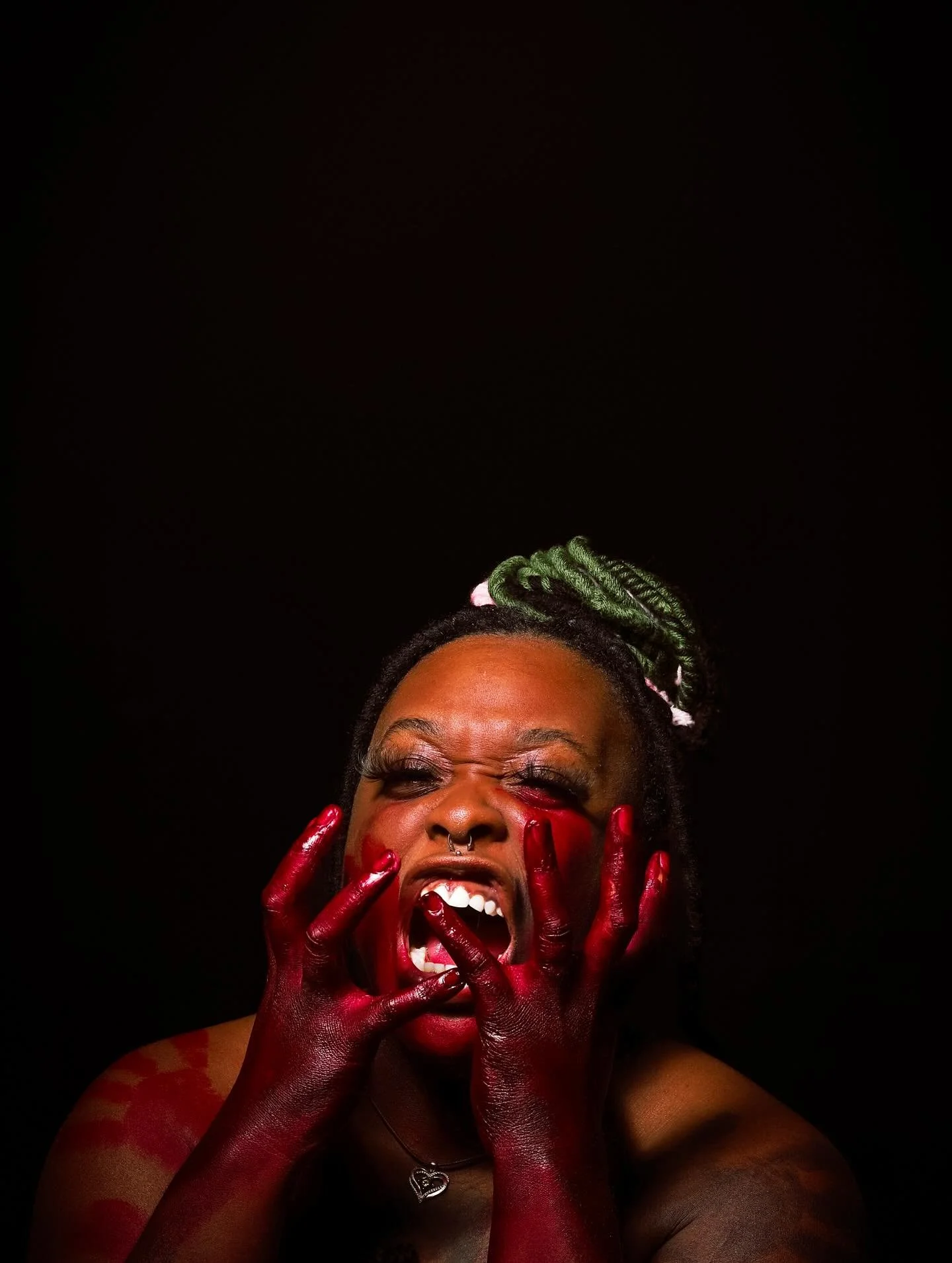 A woman with green dreadlocks and makeup, with her face contorted, hands covered in red paint covering her face, and a dark background.