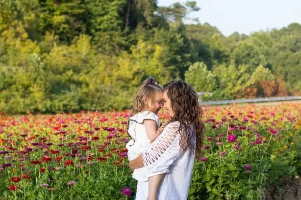 A woman holding a young girl in a field of colorful flowers with trees and a blue sky in the background.