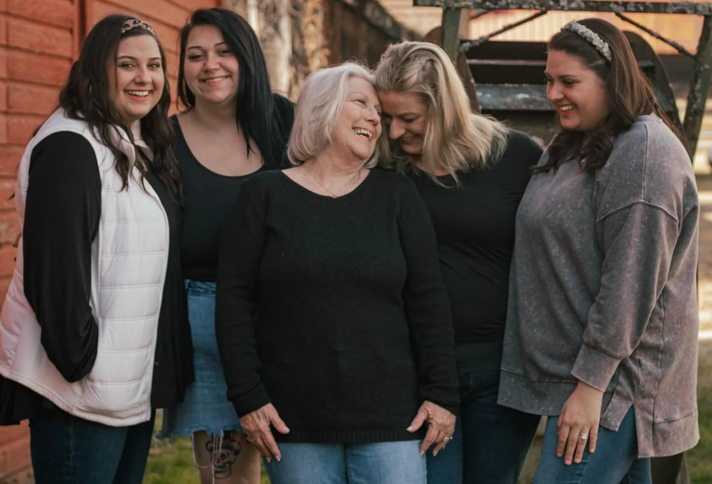 Five women, including an elderly woman, smiling and laughing together outdoors near a wooden building.