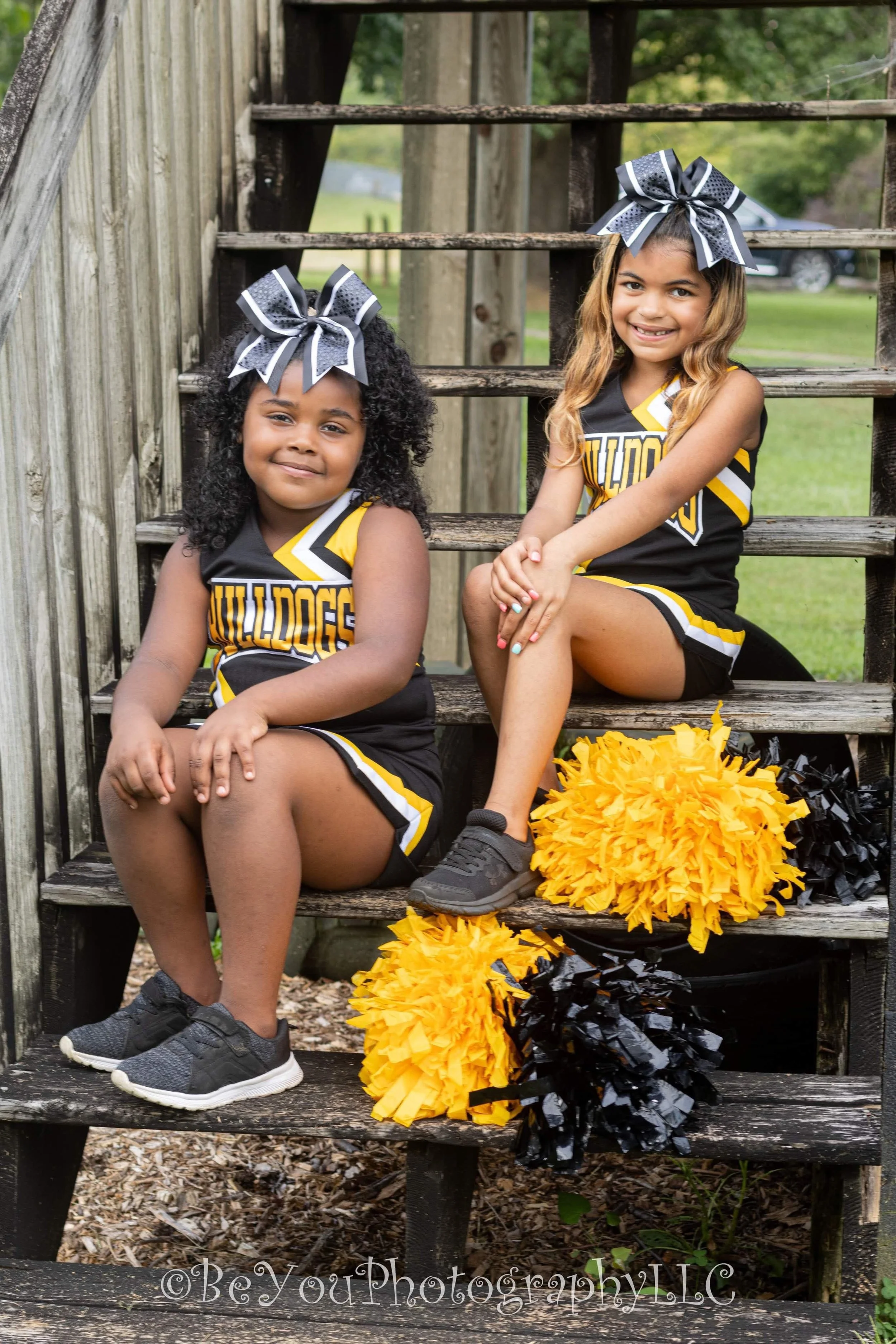 Two young cheerleaders sitting on wooden stairs outdoors, wearing black and yellow uniforms with large black and white bows on their heads, and yellow and black pom-poms beside them.